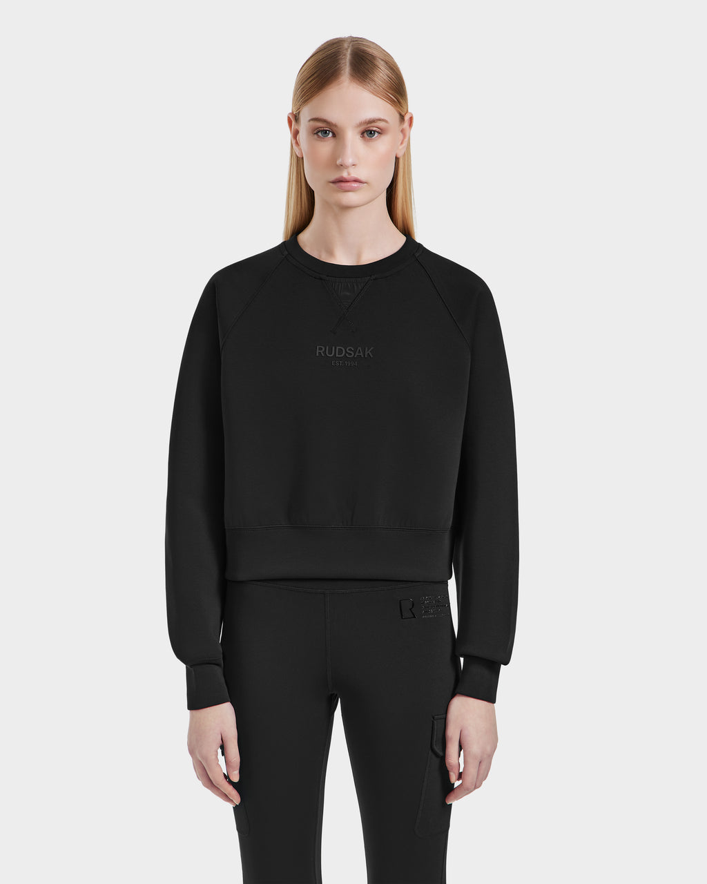 A woman with straight blonde hair wears the Rudsak BETSY - 8525087 Black crewneck sweater and black pants, standing against a plain light gray background and looking at the camera with a neutral expression.