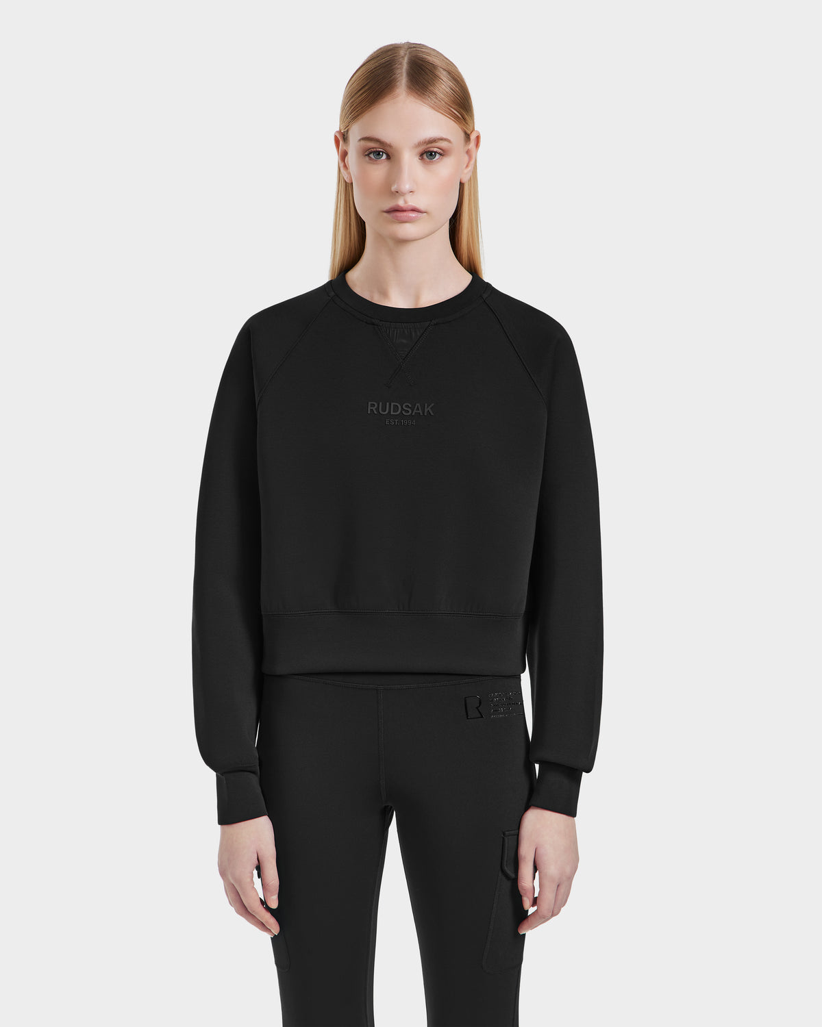 A woman with straight blonde hair wears the Rudsak BETSY - 8525087 Black crewneck sweater and black pants, standing against a plain light gray background and looking at the camera with a neutral expression.