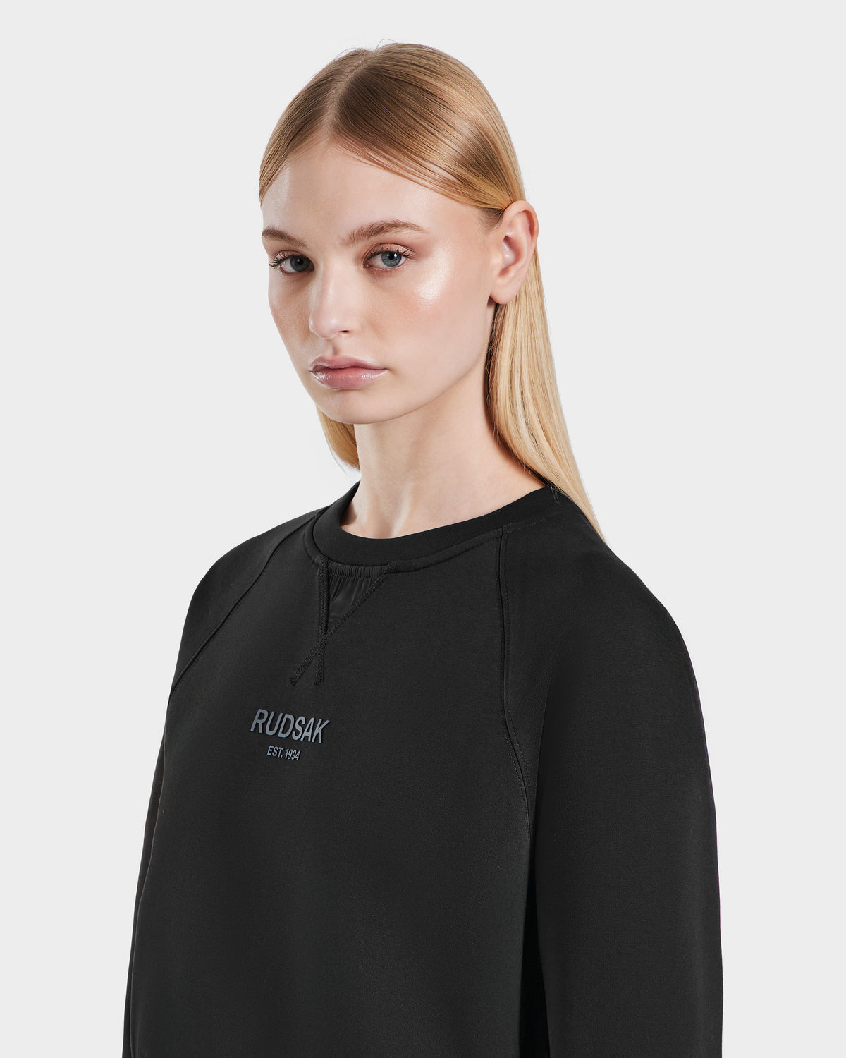A woman with straight blonde hair models the Rudsak BETSY - 8525087 womens black crop sweater featuring “RUDSAK EST 1994” on the chest. She poses against a plain light gray background, gazing slightly to her left.