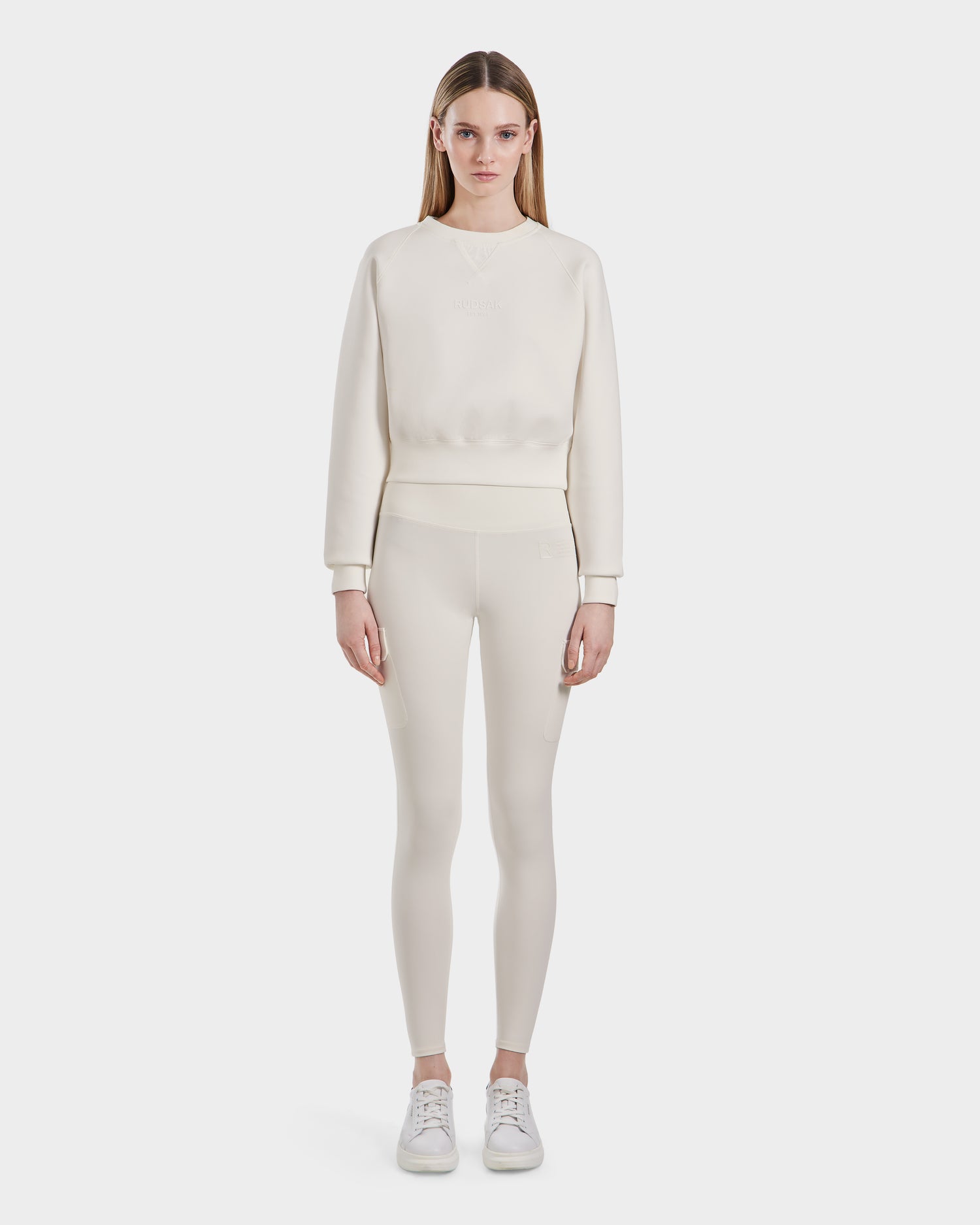 A woman faces forward against a white background, wearing the Rudsak BETSY - 8525087 Iceland off-white crewneck sweater in a premium cotton blend with matching fitted leggings and white sneakers.