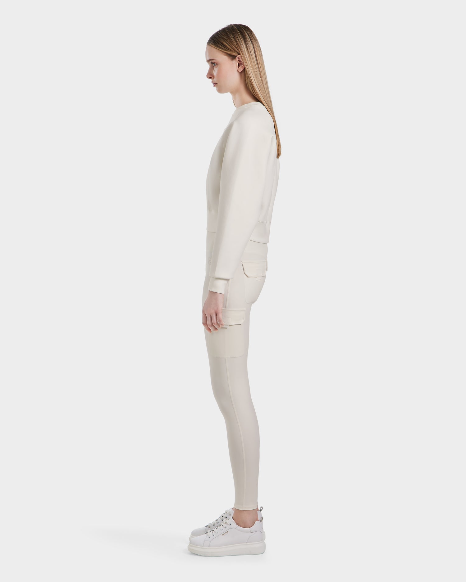 A woman stands in profile against a white background, wearing the BETSY - 8525087 Iceland sweater by Rudsak with fitted pants and platform sneakers. Her straight hair is down as she looks straight ahead.