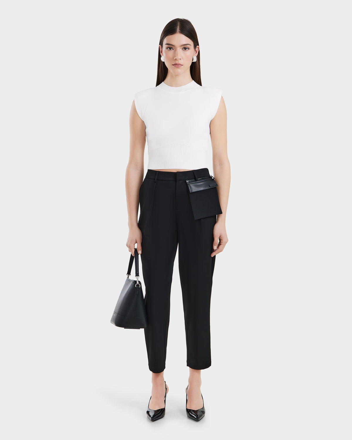 A woman with straight brown hair wears a sleeveless white crop top, tailored trousers with pocket details, black heels, and carries the BETTIE - 8525056 Black handbag by Rudsak, standing against a plain light background.
