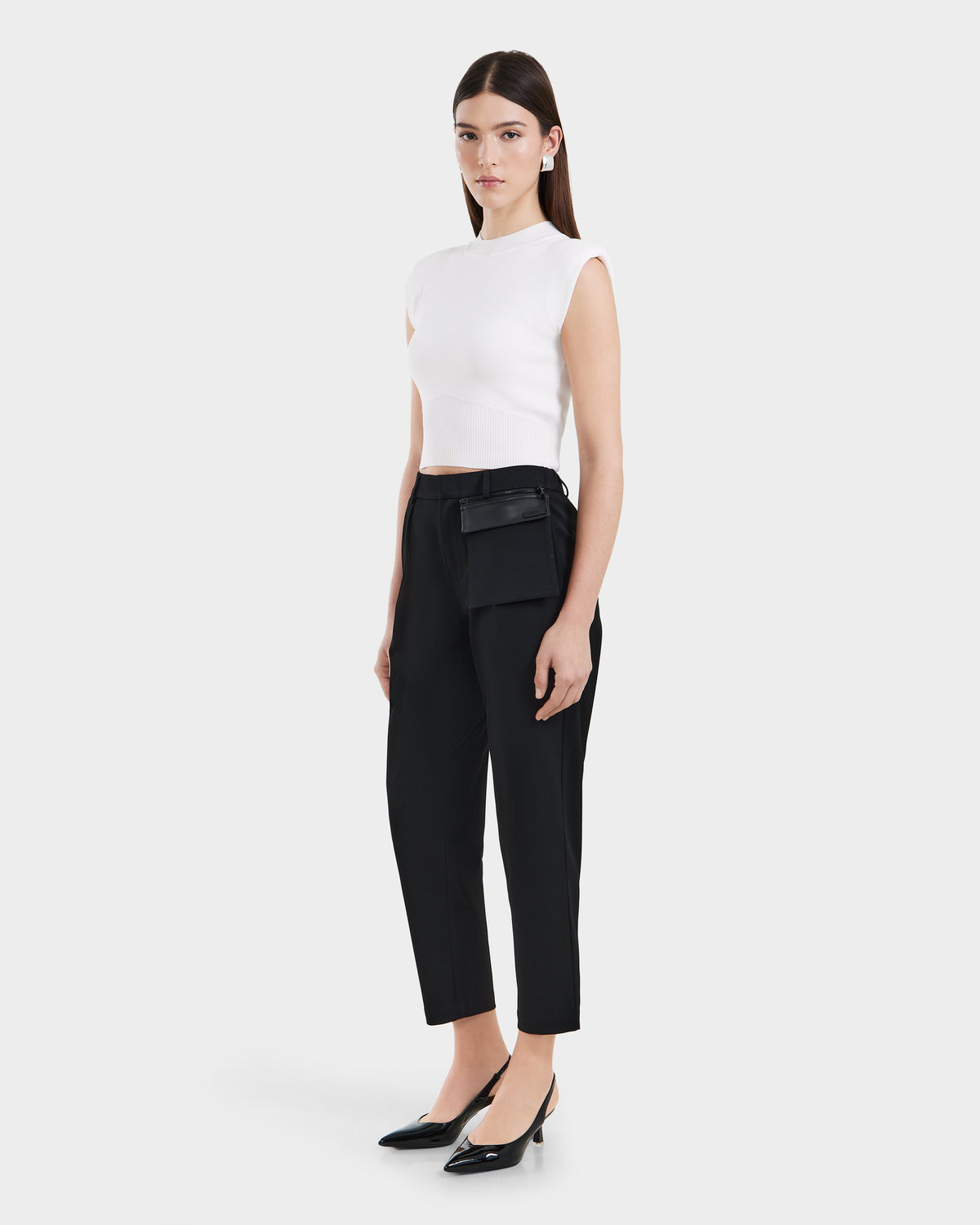 A woman with straight brown hair and a neutral expression stands against a plain background, wearing a white sleeveless top, tailored trousers, black pointed-toe heels, and the Rudsak BETTIE - 8525056 Black vegan leather belt bag for a contemporary essential look.