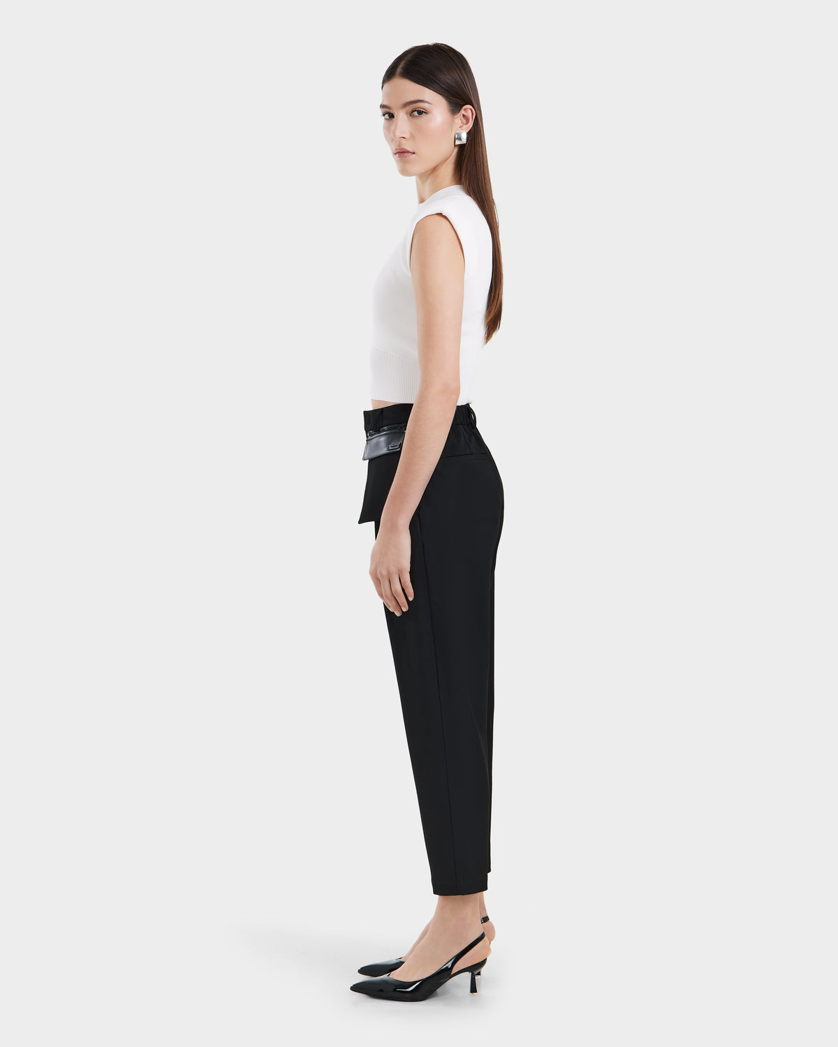 A woman stands in profile against a white background, wearing Rudsaks BETTIE - 8525056 Black pointed-toe slingback heels with black tailored trousers, a white sleeveless top, a wide black belt, and long, straight hair.