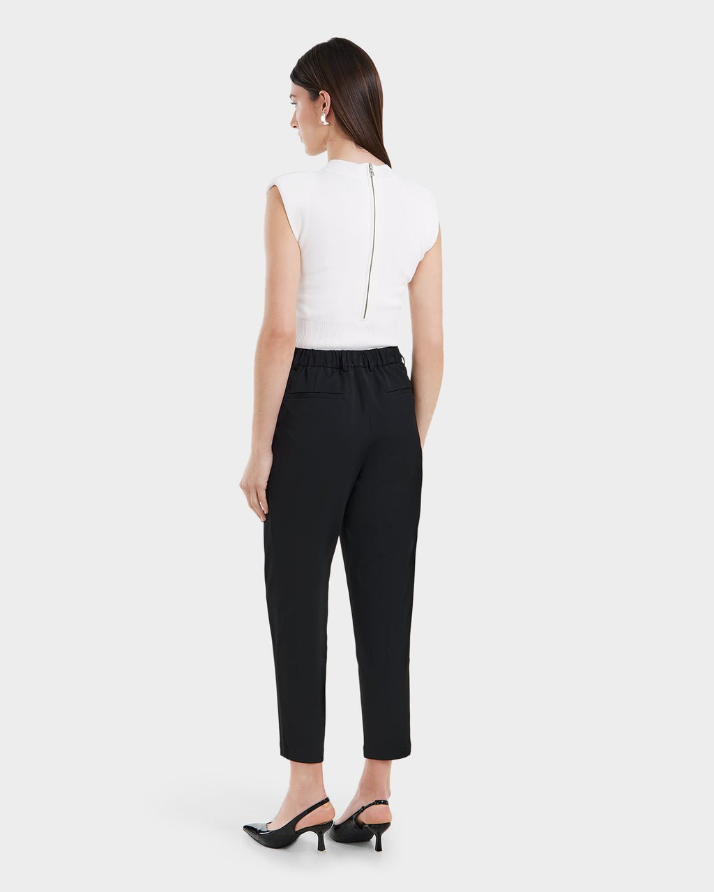 A woman with straight dark hair, facing away against a light background, wears Rudsaks BETTIE - 8525056 Black tailored trousers with a gathered waistband, paired with a sleeveless white top and black slingback heels.