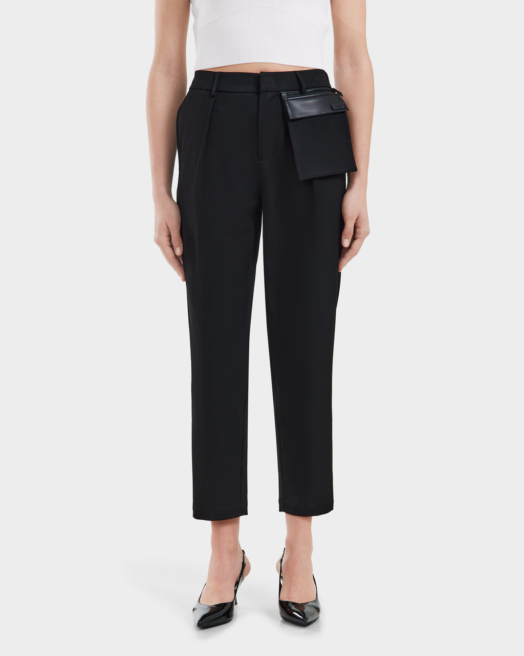 A woman models Rudsaks BETTIE - 8525056 Black pants with hip pocket detail, styled with black pointed heels and a white sleeveless crop top, set against a plain white background.
