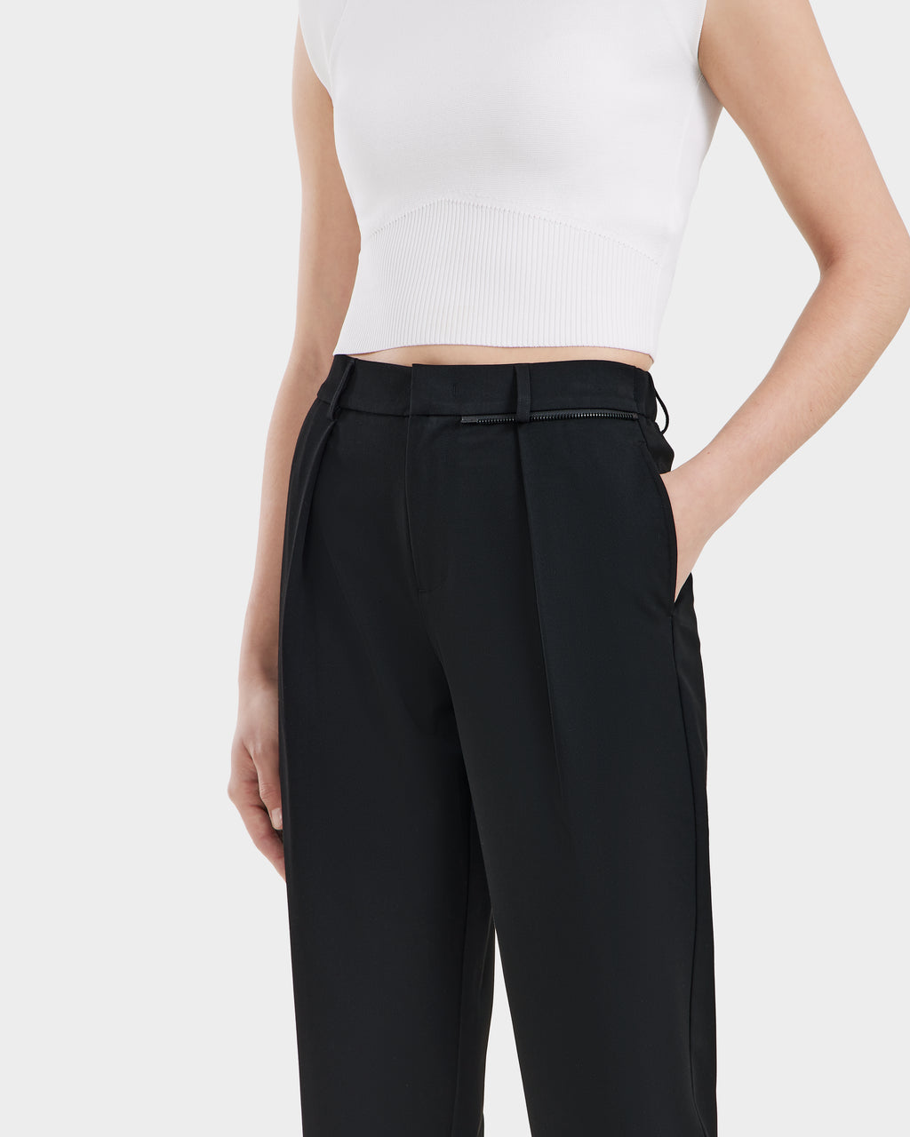 A person wears the Rudsak BETTIE - 8525056 Black tailored trousers, styled with a sleeveless white cropped top and one hand in their pocket, standing against a plain light background.