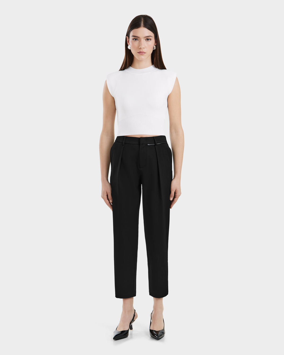 A woman faces forward in a sleeveless white crop top, high-waisted tailored trousers, and Rudsak BETTIE - 8525056 Black heels against a plain light background, embodying contemporary essential style.