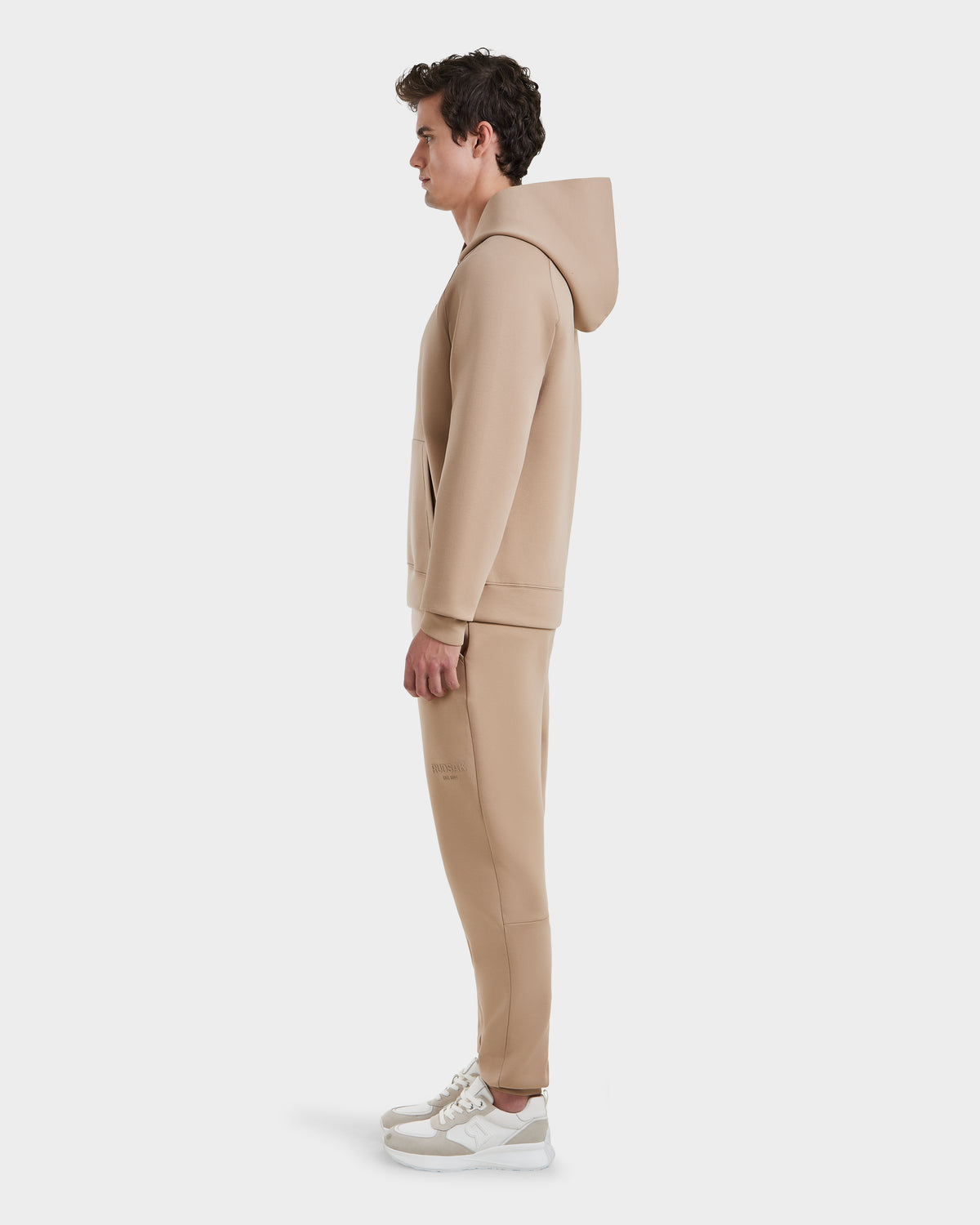 A man stands in profile against a plain white background, wearing the Rudsak BRIAR - 9525001 Almond M unisex hoodie and matching jogger pants in a premium cotton blend, paired with white sneakers for a casual look.