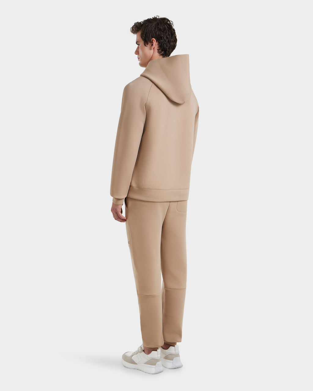 A person faces away, wearing the Rudsak BRIAR - 9525001 Almond M unisex hoodie and matching jogger pants in a premium cotton blend with white sneakers, set against a plain white background.