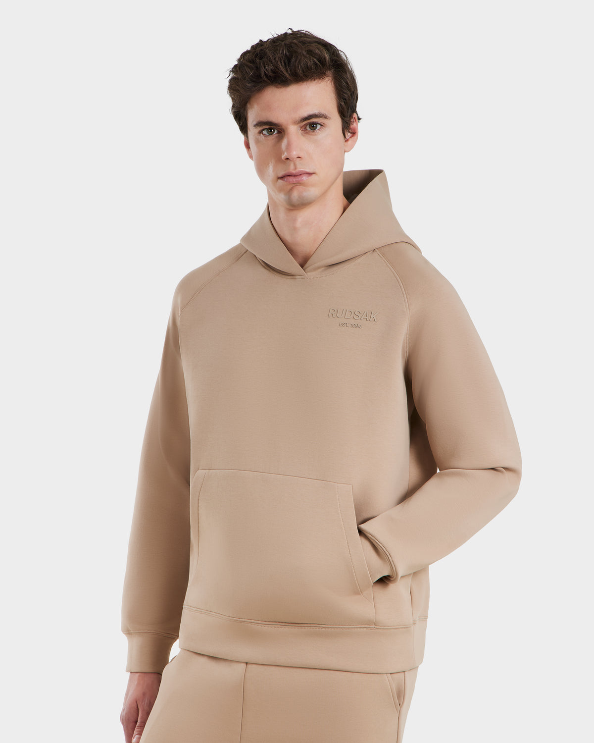 A young man models the Rudsak BRIAR - 9525001 Almond M unisex hoodie with a front pocket and matching pants, standing against a white background with a neutral expression for a relaxed, casual look.