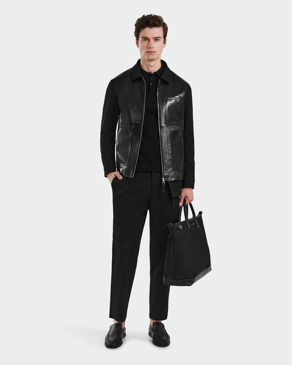 A man faces forward, dressed in the Rudsak BRYAN - 6125016 Black leather shirt jacket, black pants, and loafers. He holds a black tote bag in his left hand against a plain white background.