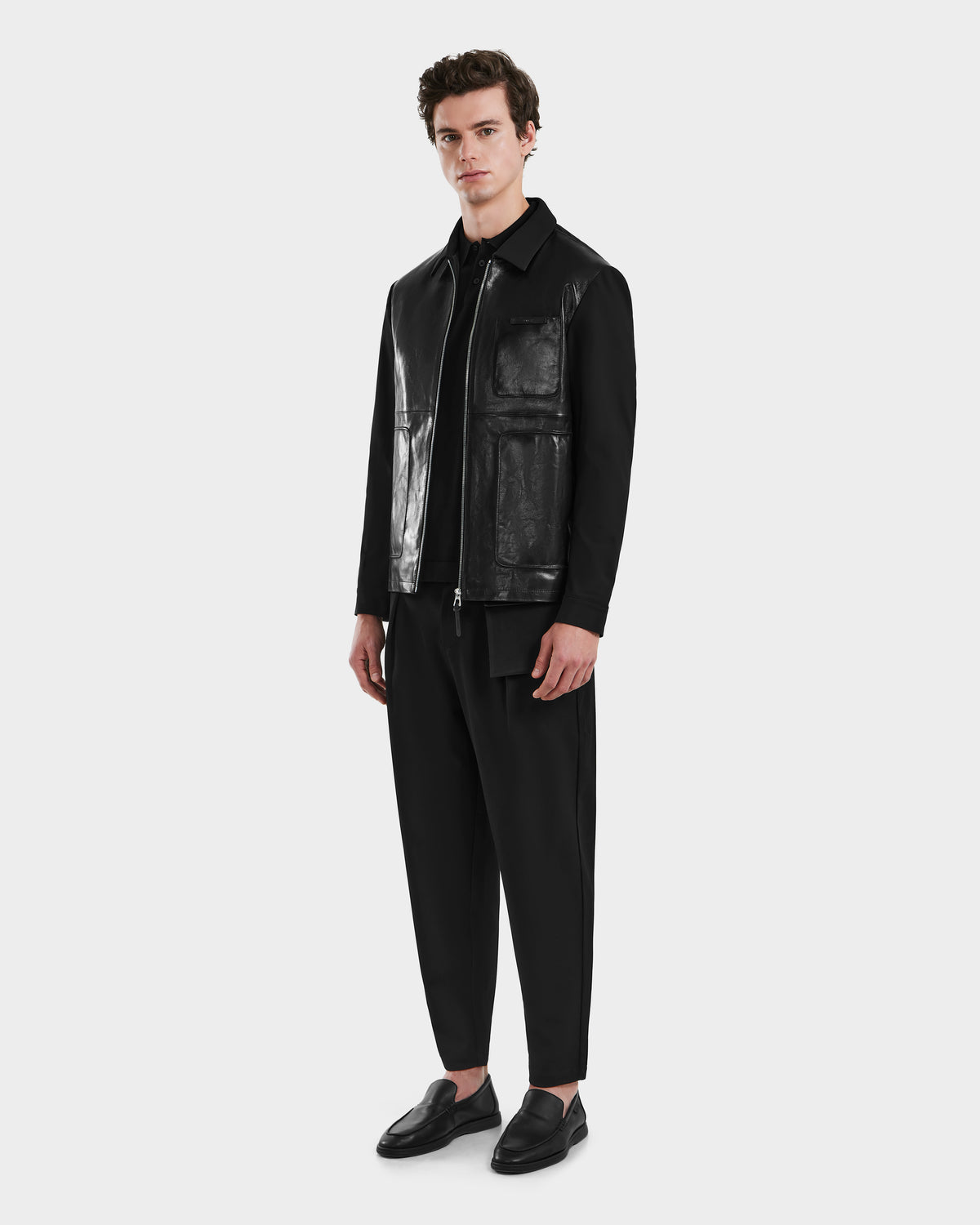 A man with short brown hair and a neutral expression stands against a plain light background, wearing the Rudsak BRYAN - 6125016 Black leather shirt jacket, black shirt and pants, and black loafers.