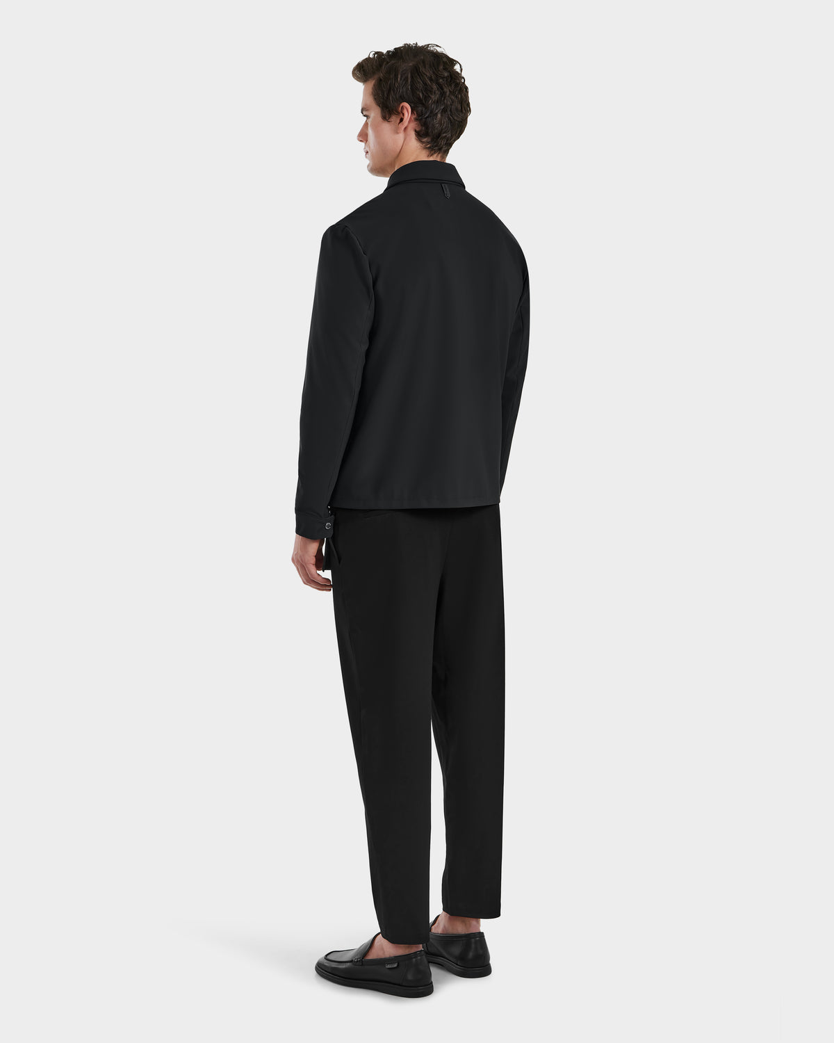A man stands facing away from the camera wearing the Rudsak BRYAN - 6125016 Black leather shirt jacket, paired with black pants and shoes, against a plain white background.