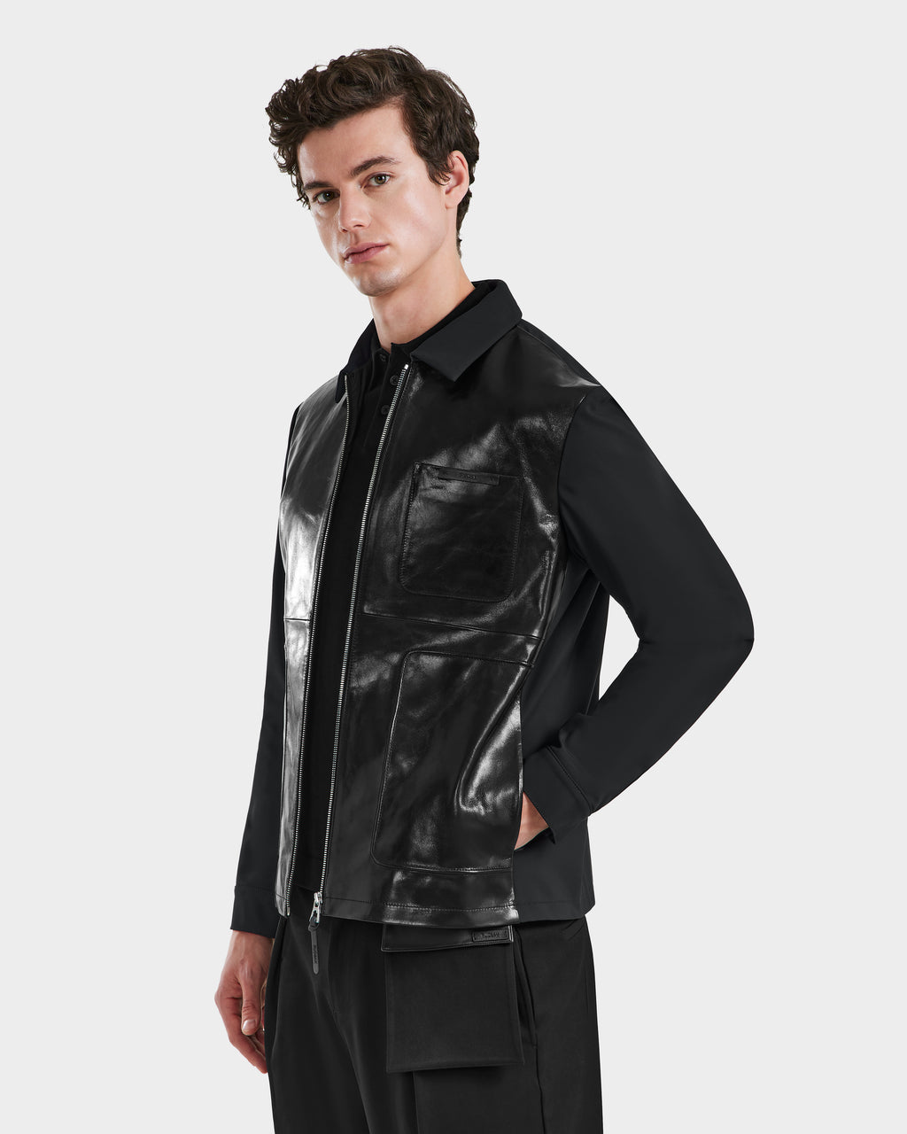 A young man with short brown hair wears the Rudsak BRYAN - 6125016 Black leather shirt jacket over a black outfit, standing confidently with one hand in his pocket against a plain light gray background.