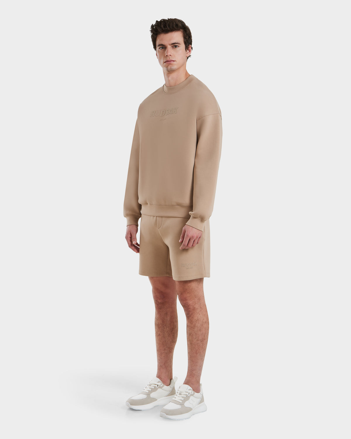 A man wears the Rudsak BRYN - 9524007 Almond M, a matching beige cotton crewneck sweatshirt and shorts set, paired with white sneakers, posing against a plain white background.