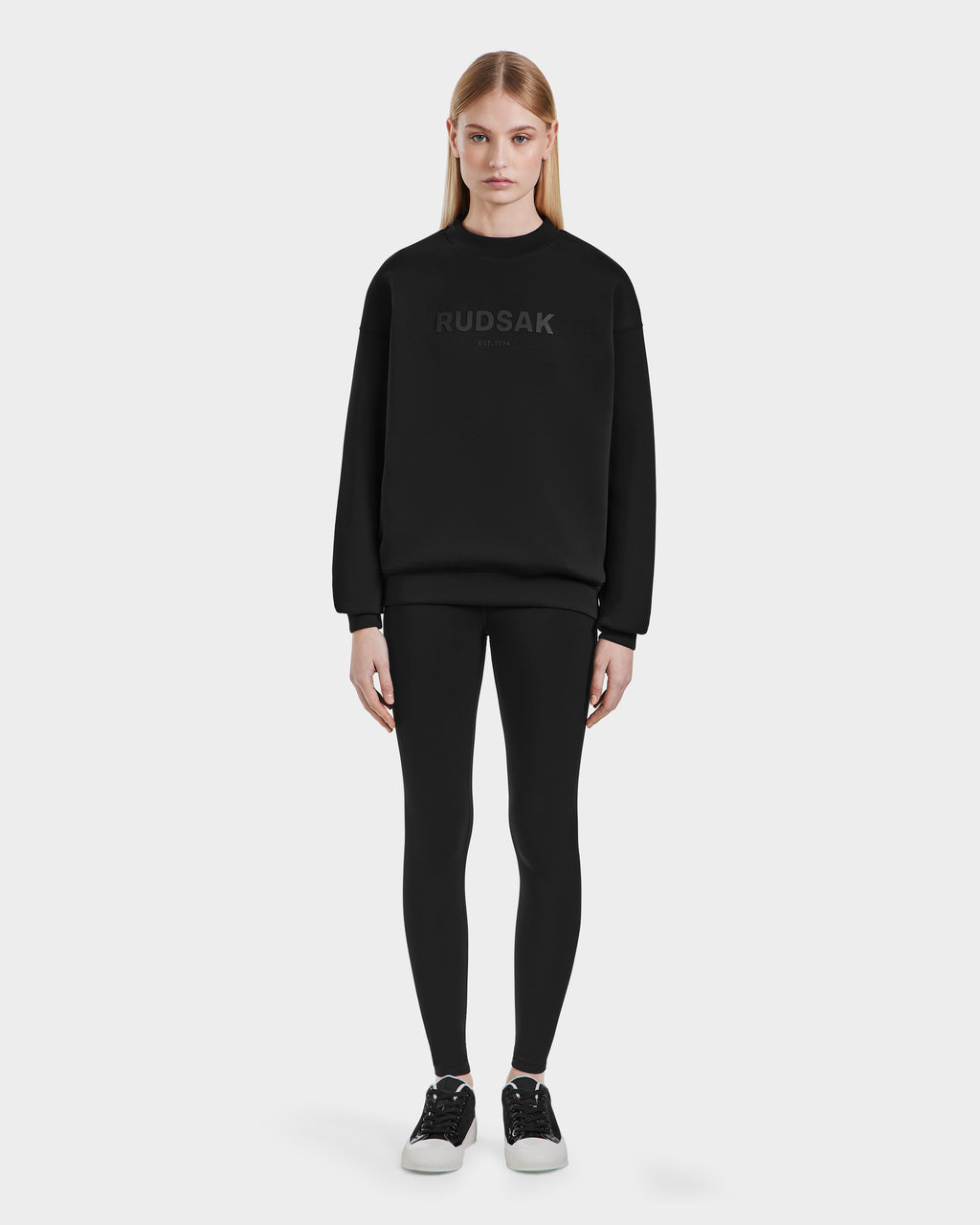 A woman faces forward wearing the Rudsak BRYN - 9524007 Black W cotton crewneck sweatshirt, paired with black leggings and black sneakers with white soles, standing against a plain white background to showcase an everyday look.
