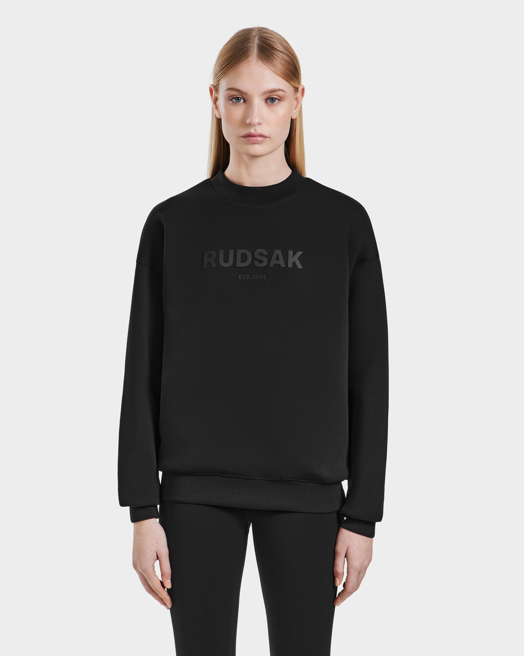 A person with straight blonde hair models the Rudsak BRYN - 9524007 Black W cotton crewneck sweatshirt, featuring RUDSAK EST. 1994 on the front, in a genderless everyday fit, against a plain white background.