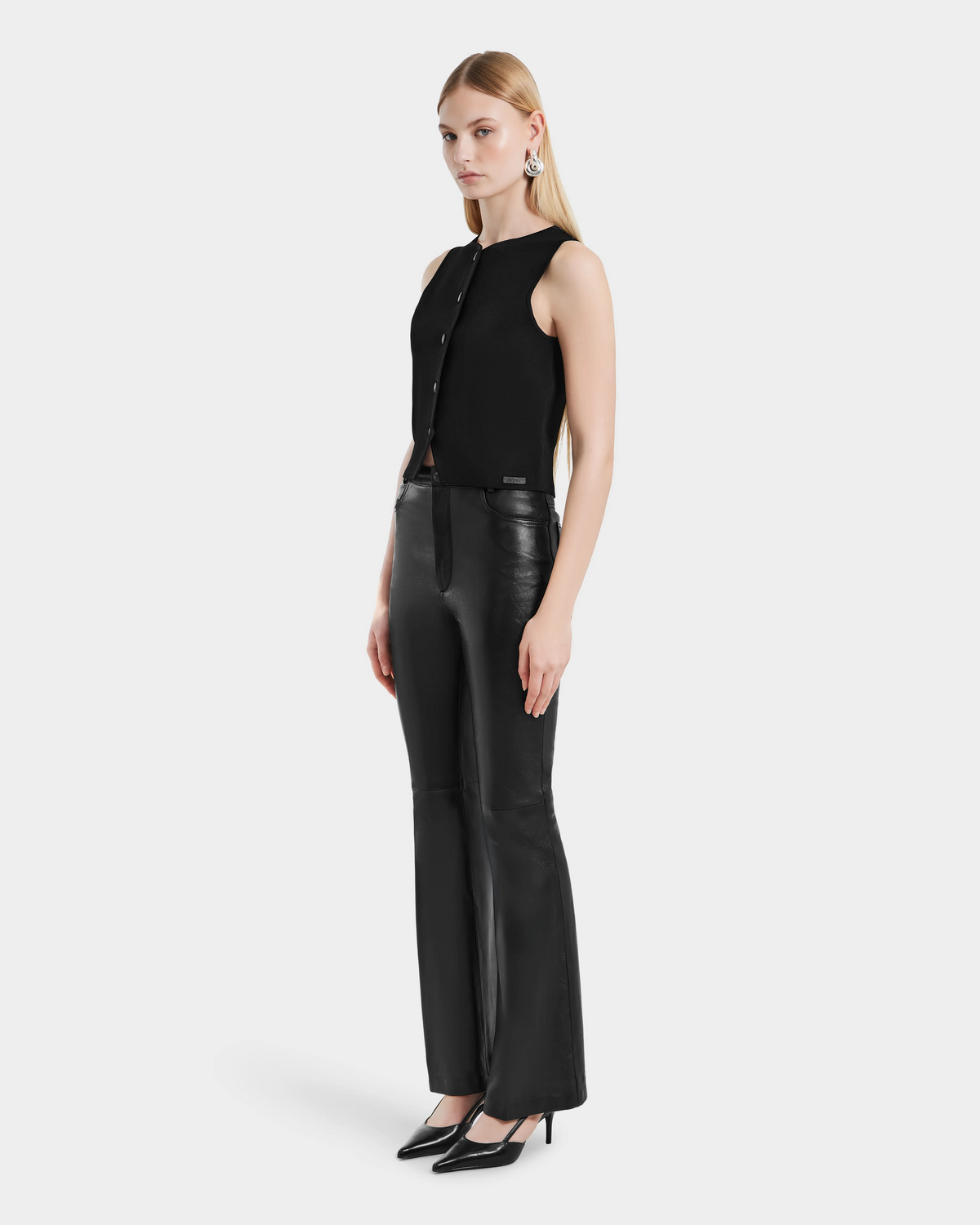 A woman with straight blonde hair and a neutral expression models the Rudsak CARISSA - 8524545 Black sleeveless buttoned top, styled with womens leather pants, black heels, and statement silver earrings against a plain white background.