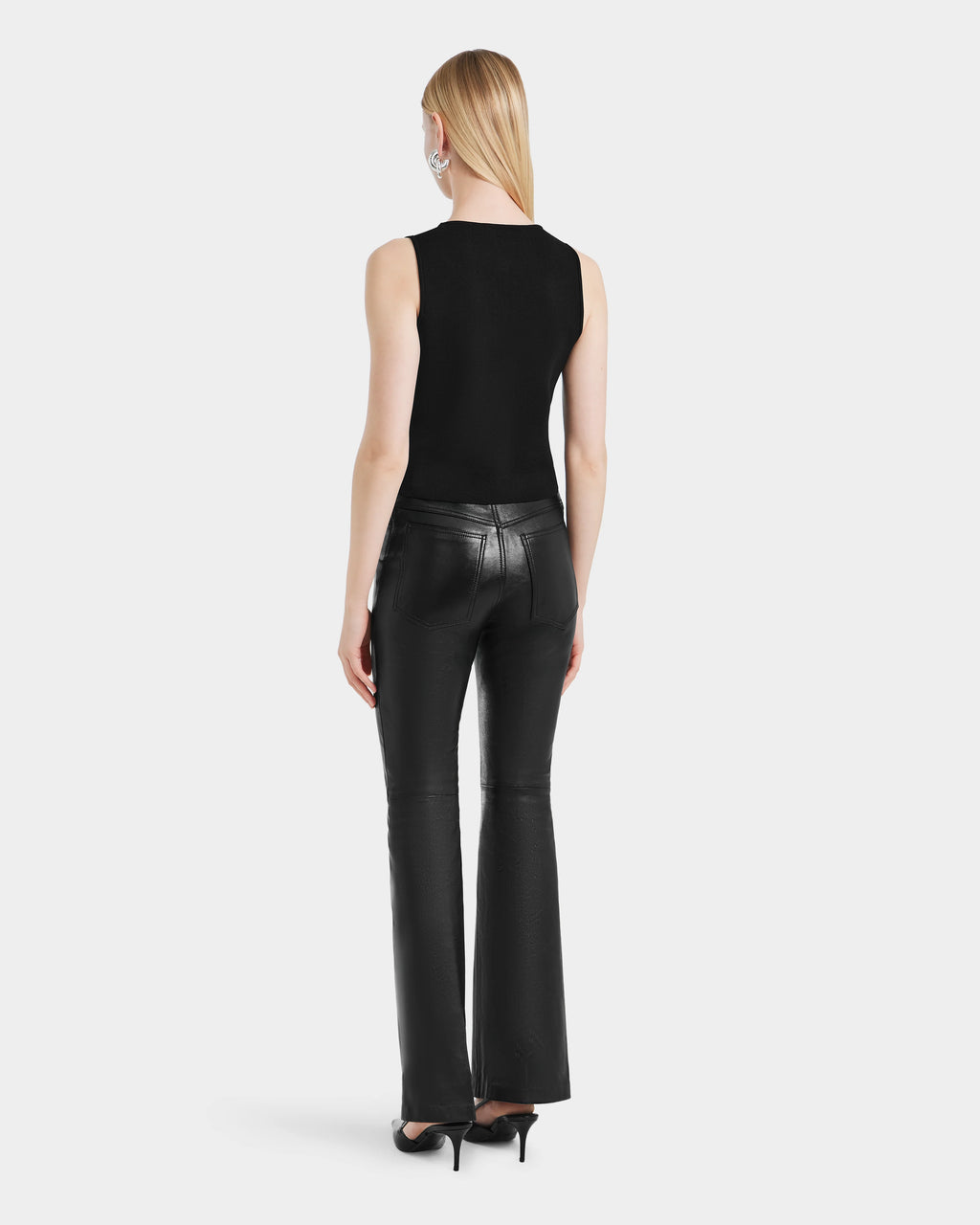 A person with straight blonde hair stands facing away against a plain light background, wearing the Rudsak CARISSA - 8524545 Black sleeveless top and womens flared leather pants, paired with black high-heeled sandals.