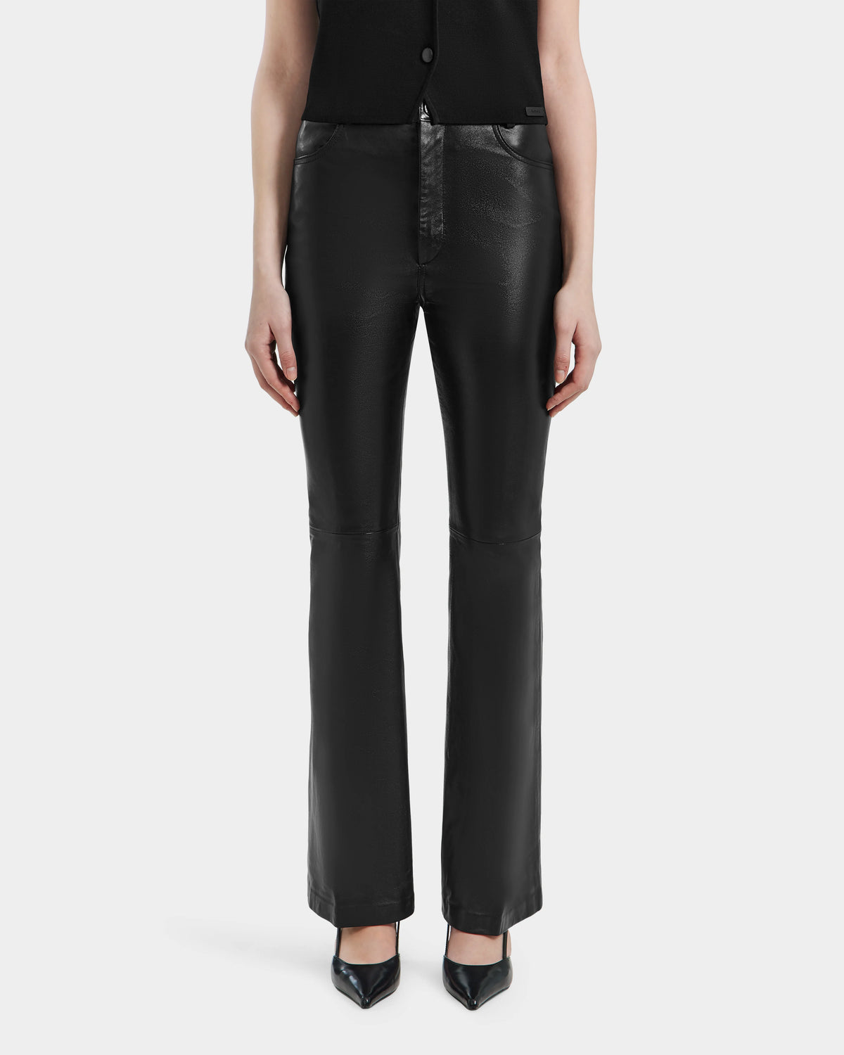 A person stands facing forward in Rudsak’s CARISSA - 8524545 Black leather pants with black pointed-toe heels. Only the lower torso, legs, and arms are shown against a plain white background.