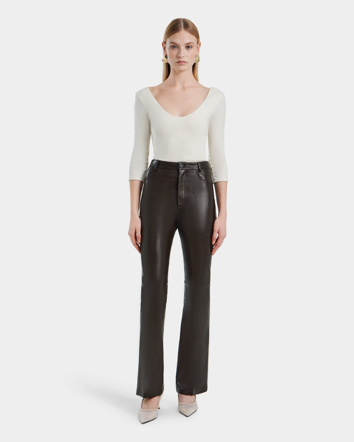 A woman faces forward against a white background, wearing Rudsaks CARISSA - 8524545 Espresso high-waisted womens leather pants, a white scoop neck top, and light gray pointed-toe heels.