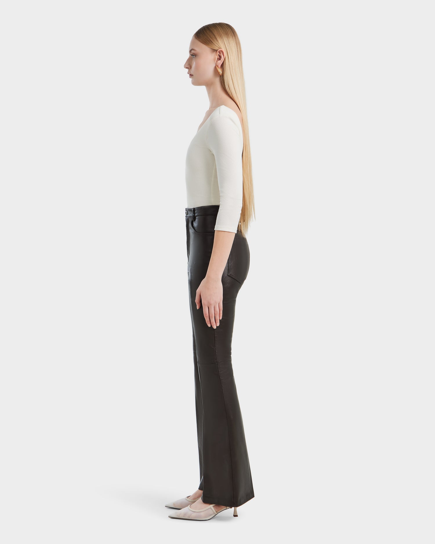 A woman with long straight blonde hair stands in profile against a white background, wearing Rudsaks CARISSA - 8524545 Espresso high-waisted black womens leather pants, a white fitted top, and beige pointed-toe heels.