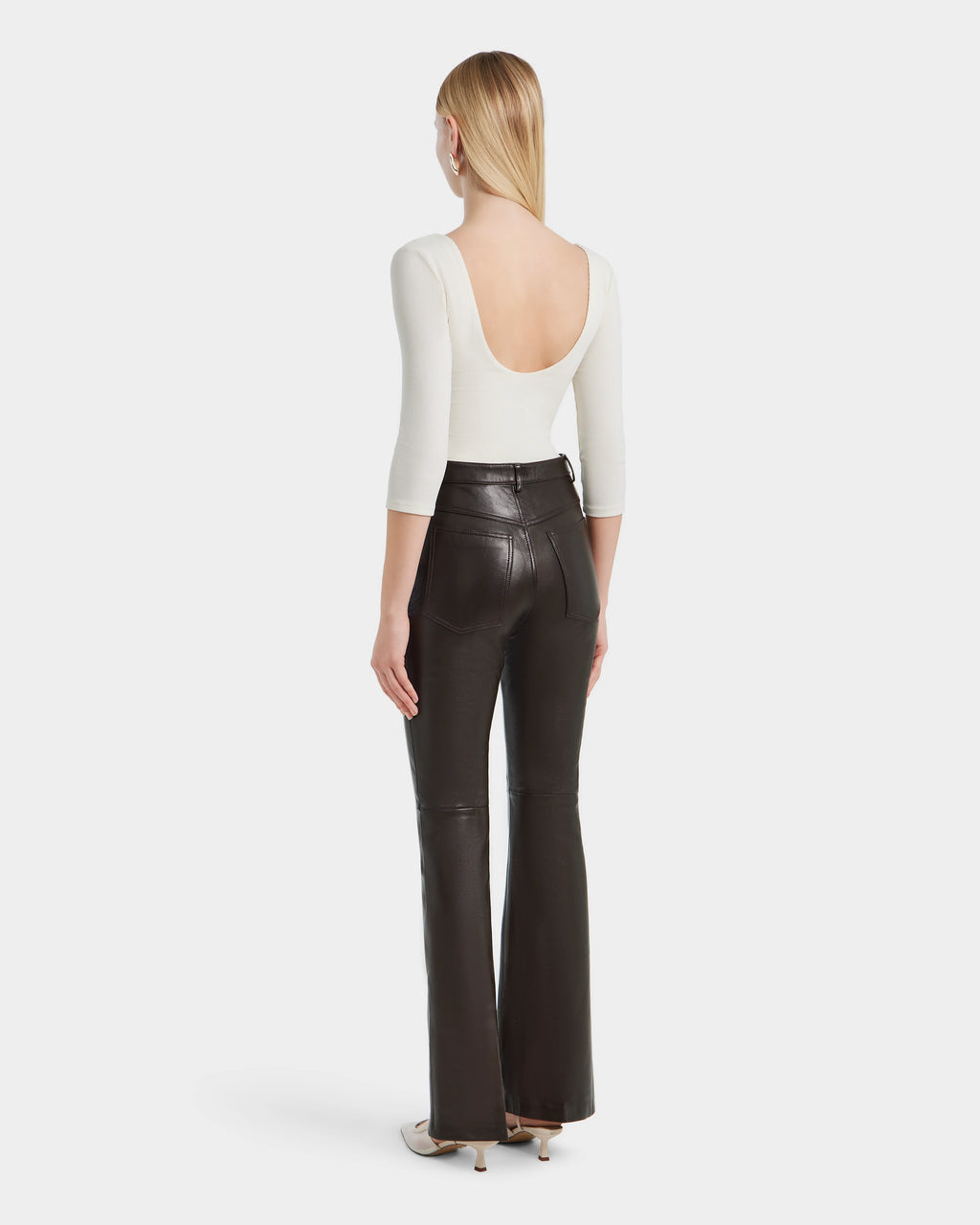 A woman faces away in Rudsaks CARISSA - 8524545 Espresso high-waisted flare leather pants, paired with a white scoop-back three-quarter sleeve top and heeled shoes. Her straight blonde hair flows loose against a plain white background.