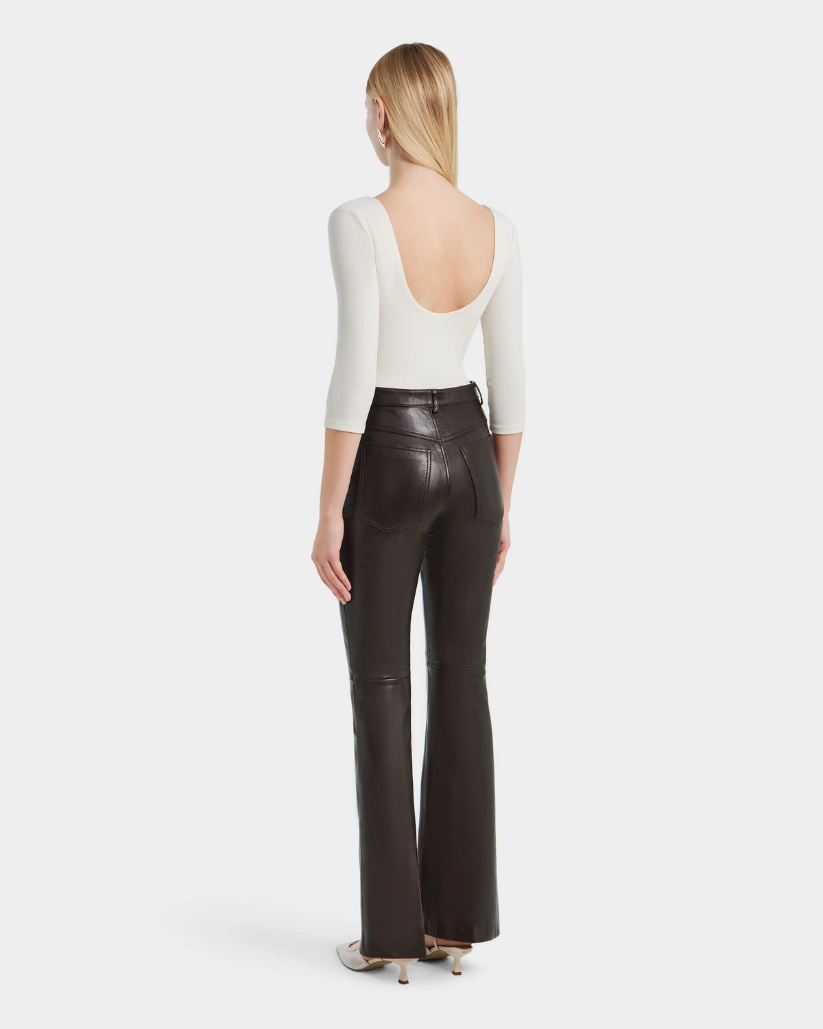 A woman faces away in Rudsaks CARISSA - 8524545 Espresso high-waisted flare leather pants, paired with a white scoop-back three-quarter sleeve top and heeled shoes. Her straight blonde hair flows loose against a plain white background.