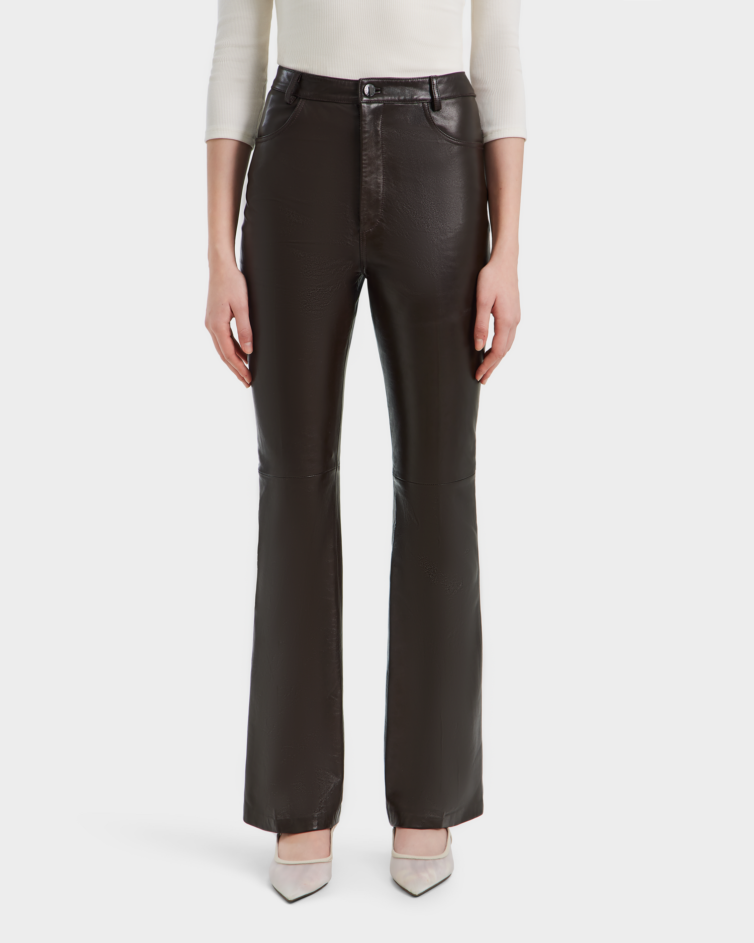 Wearing Rudsak’s CARISSA - 8524545 Espresso leather flare pants with a white long-sleeve top and light pointed-toe heels, the person stands against a plain white background; image cropped below shoulders for a chic, modern vibe.