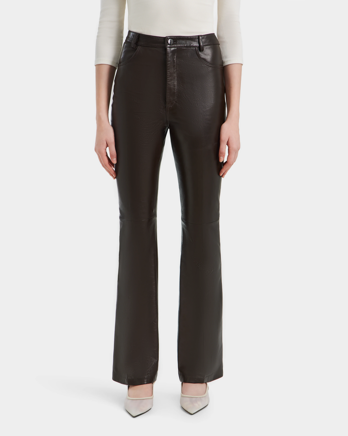 A person wears Rudsak’s CARISSA - 8524545 Espresso high-waisted womens leather pants with a straight-leg cut, paired with a white long-sleeve top and pointed-toe beige heels, shown from the shoulders down on a white background.