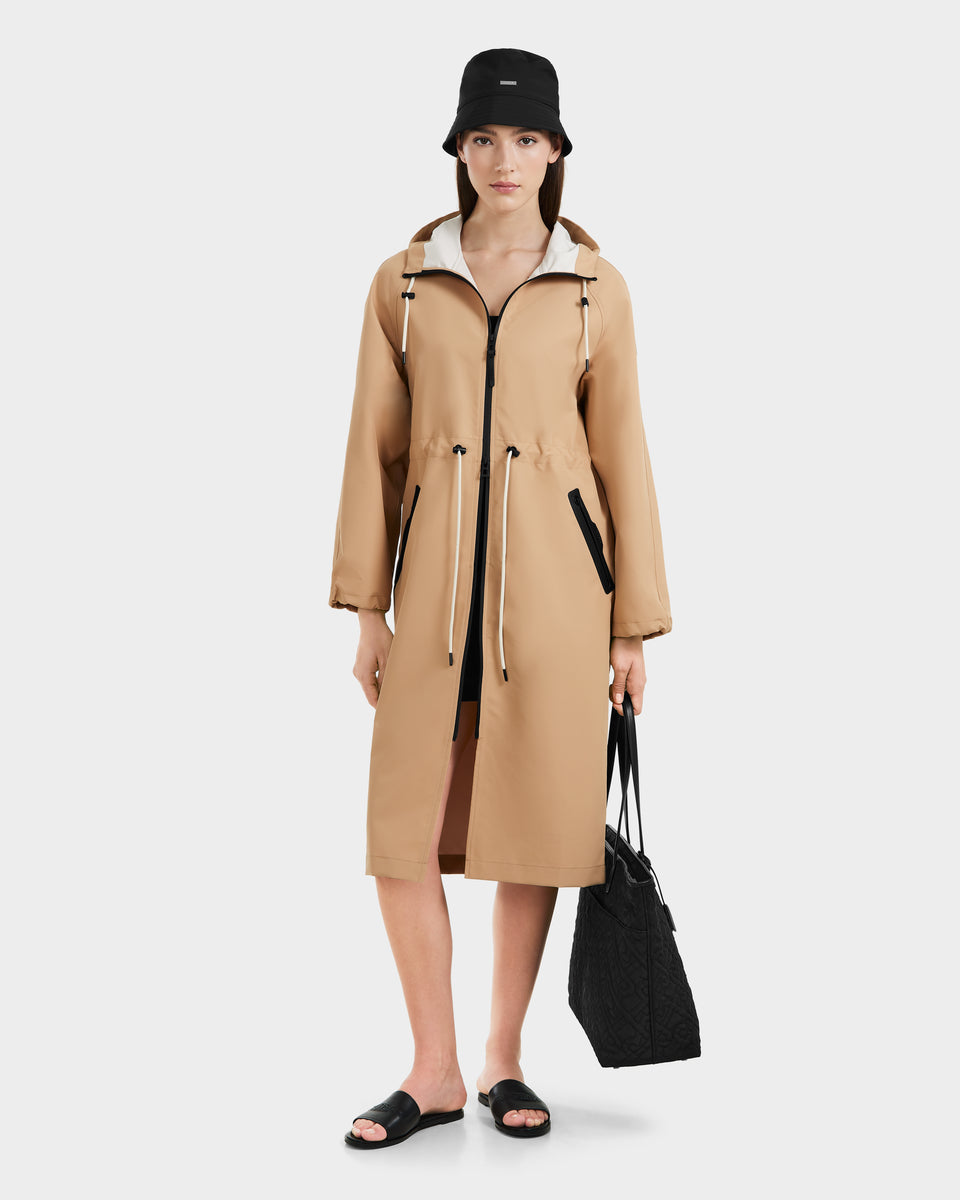 A woman wearing the Rudsak CARLA - 8125008 Almond long raincoat faces forward against a plain white background, styled with a black bucket hat, black slide sandals, and holding a black quilted tote bag.