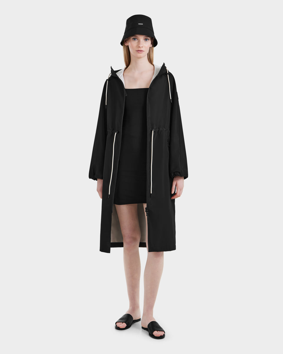 A woman faces forward with a neutral expression, wearing Rudsak’s CARLA - 8125008 Black women’s long raincoat with white drawstrings and vegan leather details, paired with a black bucket hat, dress, and slides against a white background.