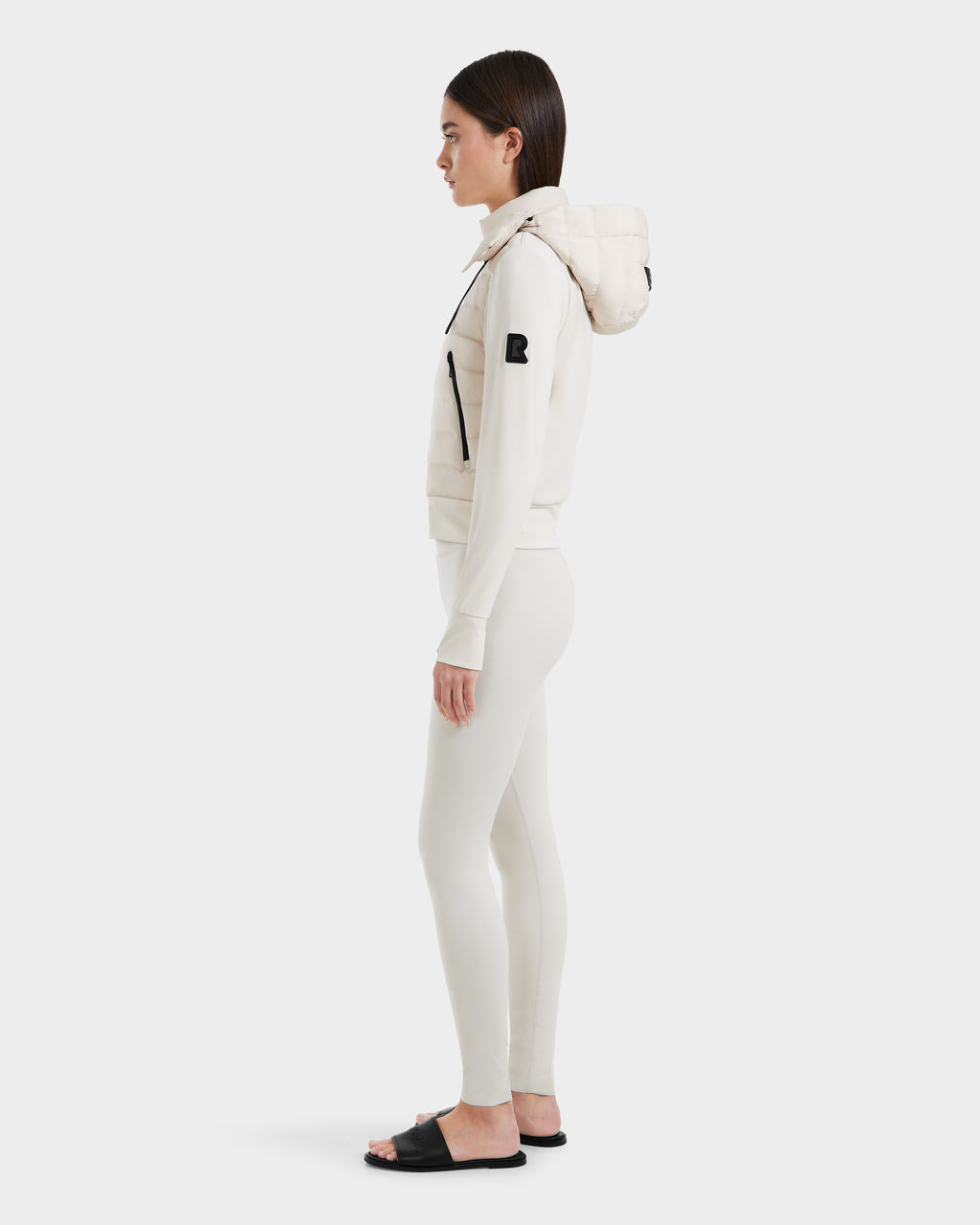 A woman in profile wears the Rudsak CASSA - 8125057 Vanilla cream hoodie with quilted hood, matching leggings, and black slide sandals against a white background—perfect to pair with a women’s transitional jacket for changing seasons.