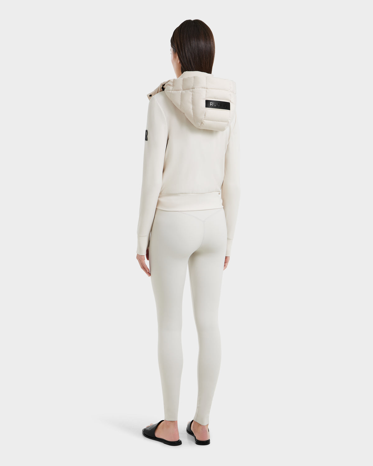 A person with straight dark hair faces away, wearing Rudsaks CASSA - 8125057 Vanilla womens transitional jacket with the hood up, fitted cream leggings, and black slide sandals against a plain white background.