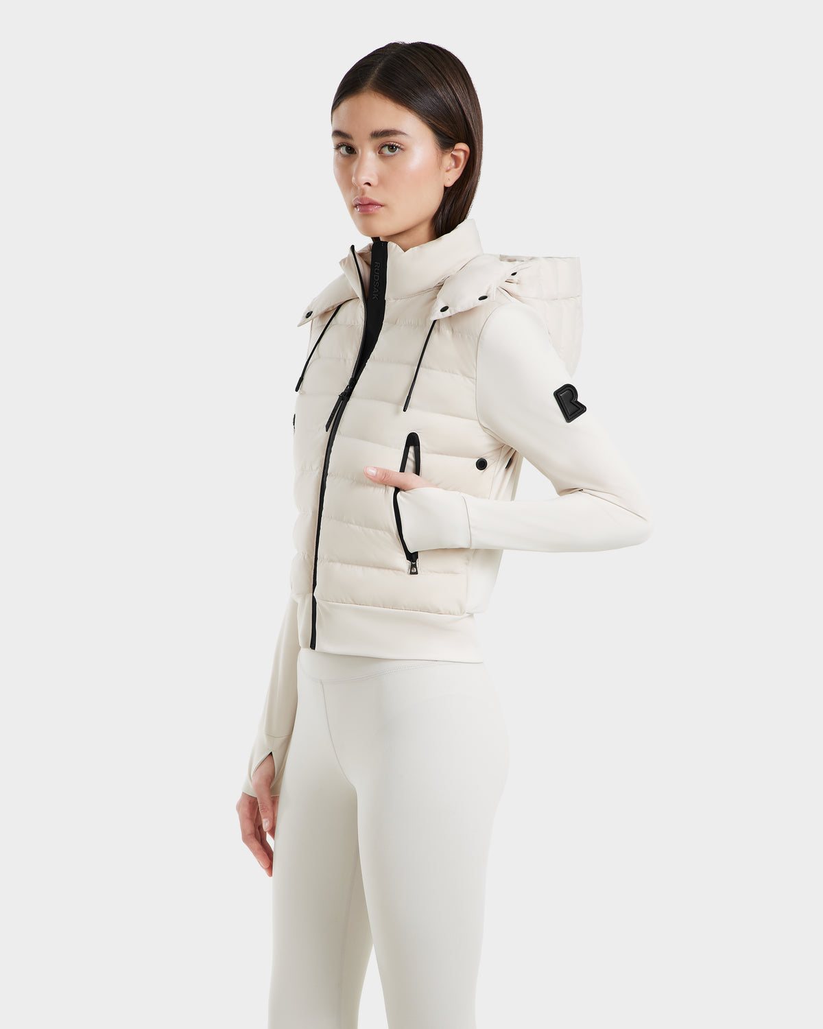 A woman wears the Rudsak CASSA - 8125057 Vanilla transitional jacket with a hood and zipper over a fitted top and leggings, standing sideways and looking at the camera against a plain white background.