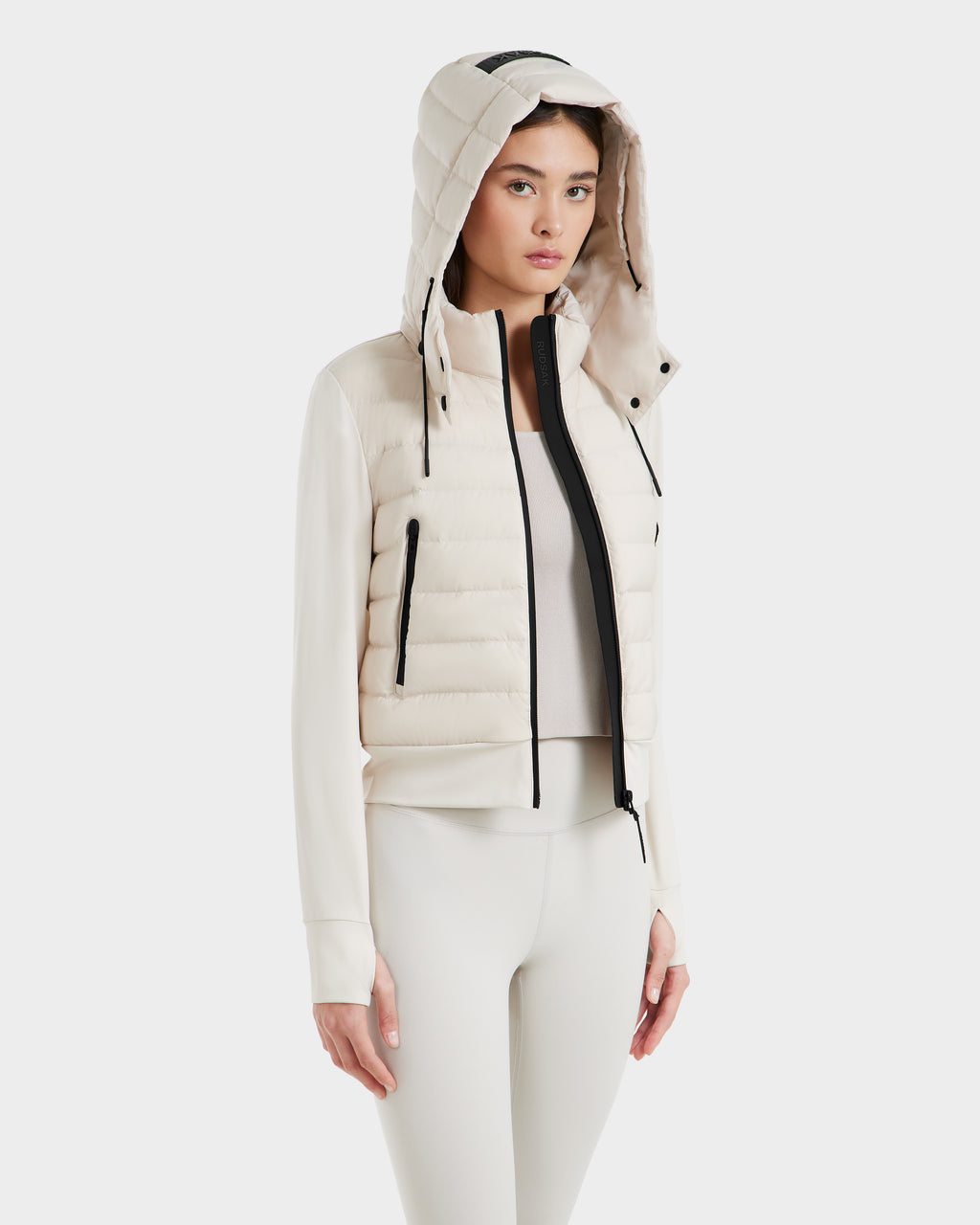 A woman models the Rudsak CASSA - 8125057 Vanilla, a lightweight, light-colored hooded puffer vest, over a fitted long-sleeve top and matching leggings against a plain white background.