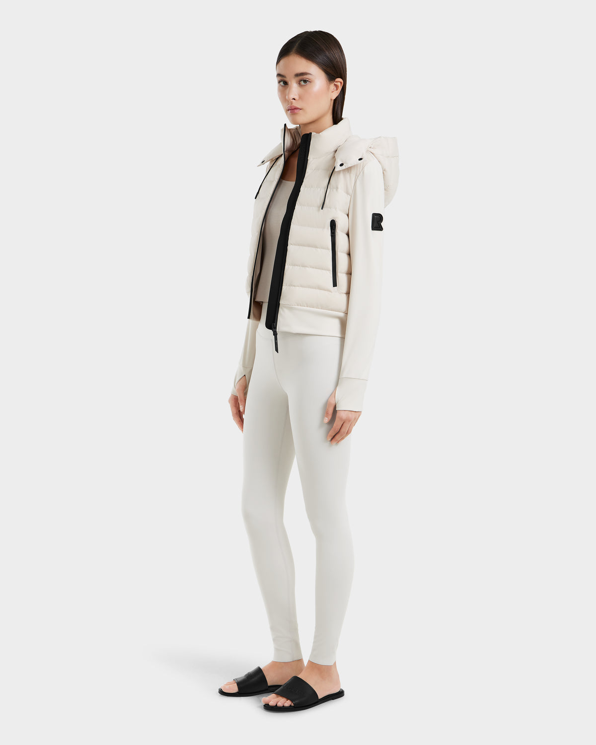 A woman with straight brown hair and a neutral expression models the Rudsak CASSA - 8125057 Vanilla, a white lightweight cropped jacket, over white leggings and a beige top with black slide sandals against a plain white background.