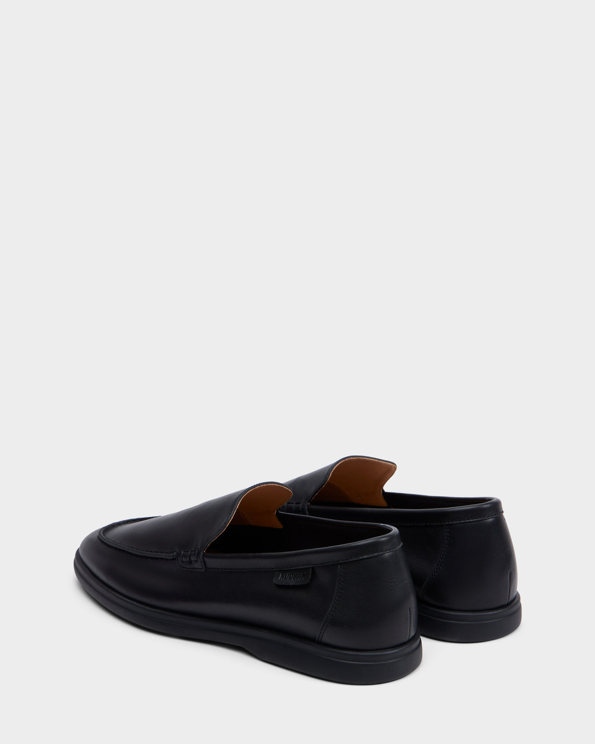 The Rudsak CHANNING - 6225005 Black men’s leather slip-on loafers are displayed on a light gray background, with one shoe positioned forward to showcase side and back views—ideal for lovers of premium footwear.