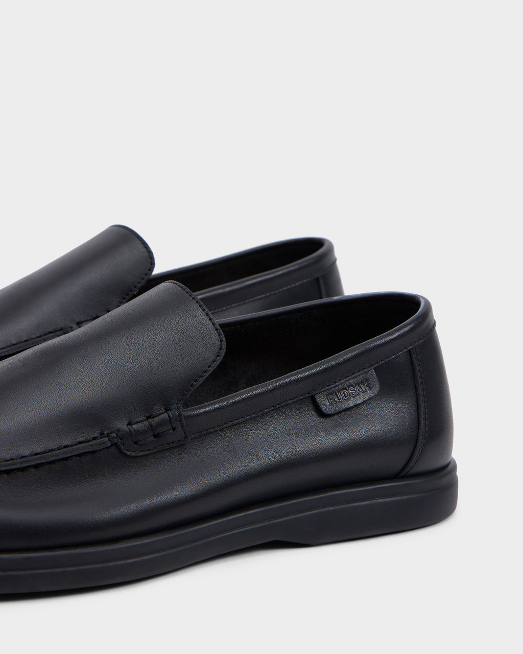 Close-up of the Rudsak CHANNING - 6225005 men’s black leather loafer with stitched seam detail, featuring a subtle side label, displayed on a light gray background—highlighting sophisticated, premium footwear.