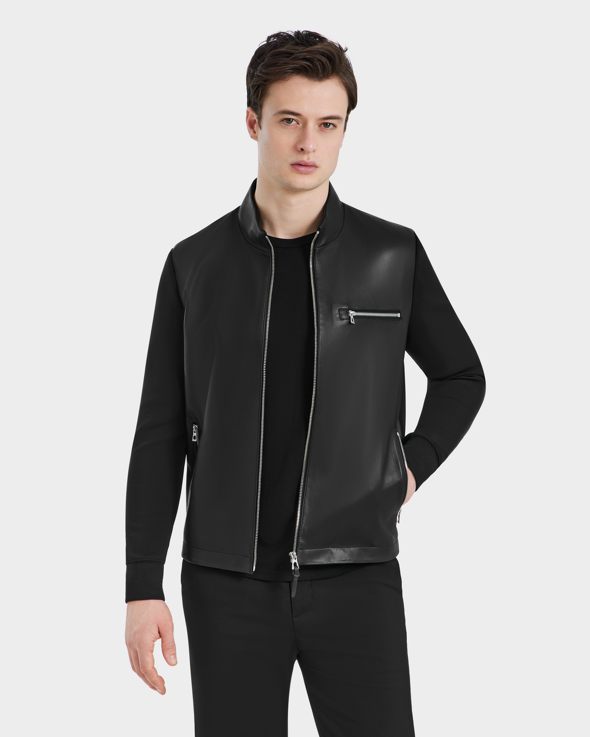A young man stands against a plain light background, wearing the Rudsak CLIFTON - 6125021 Black leather jacket over a black shirt and pants, one hand in his pocket and a neutral expression showcasing the jackets refined craftsmanship.