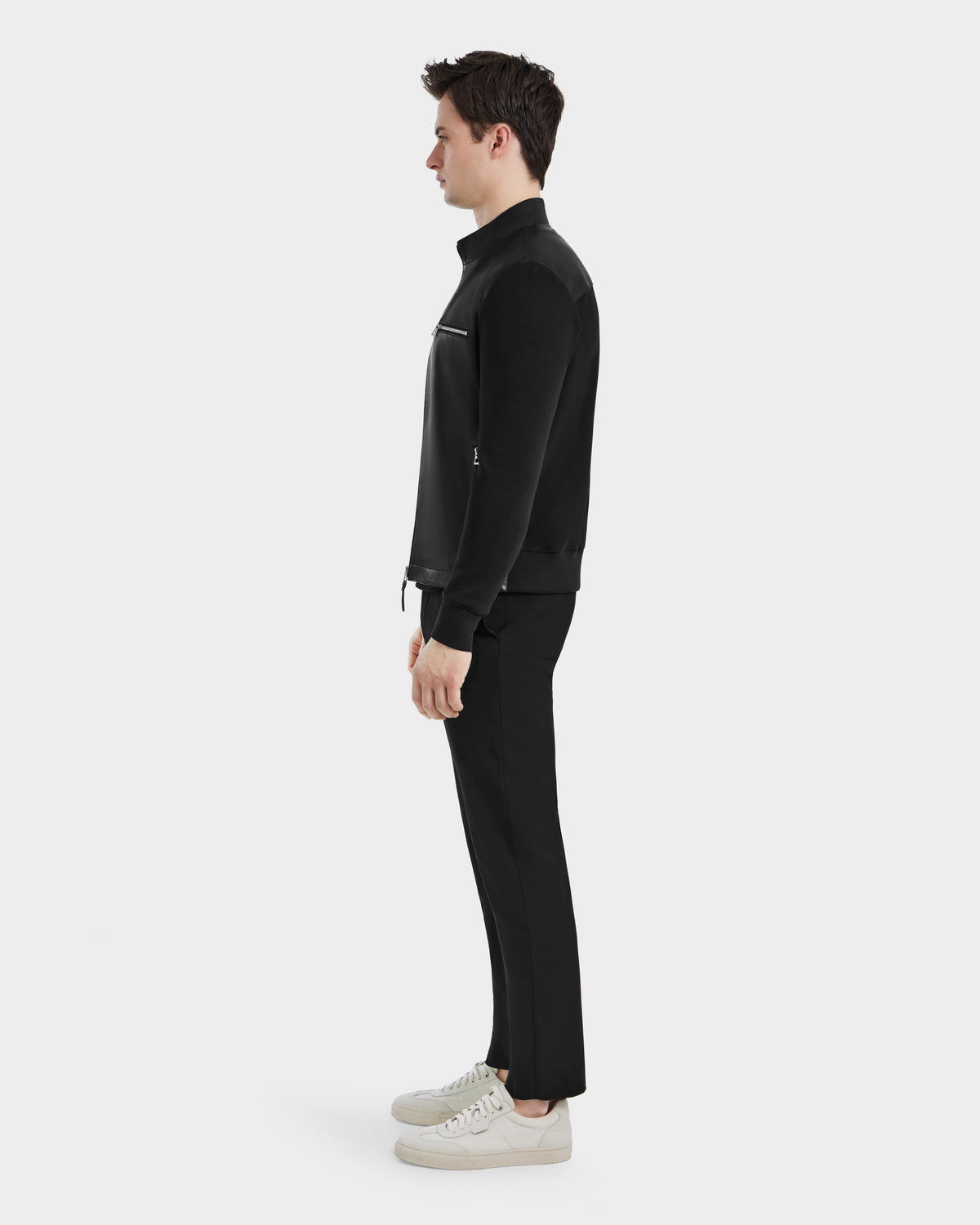 A man stands in profile facing left, wearing the Rudsak CLIFTON - 6125021 Black zip-up jacket, black pants, and white sneakers against a plain white background.