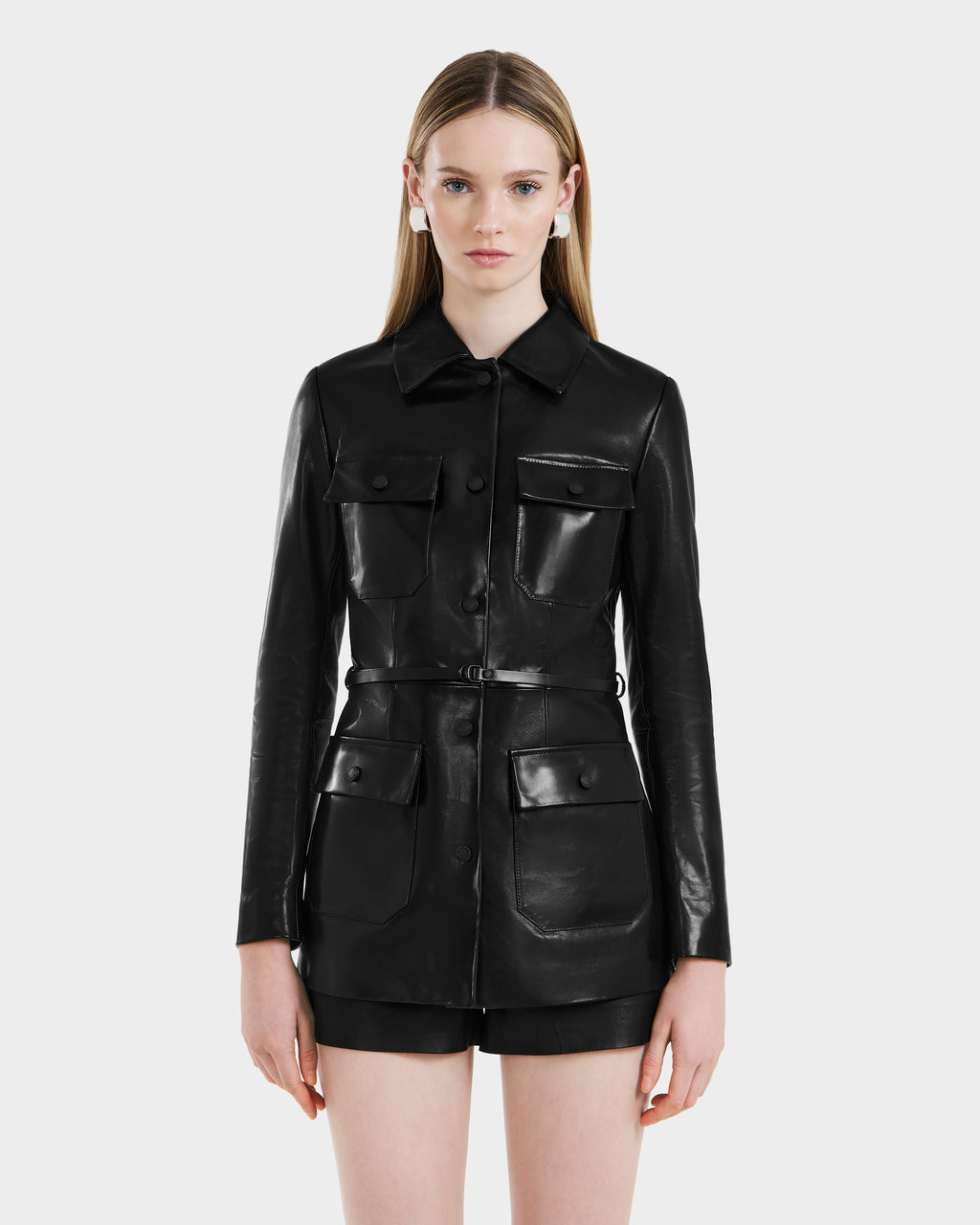 A woman with straight, long blonde hair models the Rudsak DANICA - 8125025 Black leather jacket, featuring four front pockets and buttons, paired with black shorts against a plain white background.