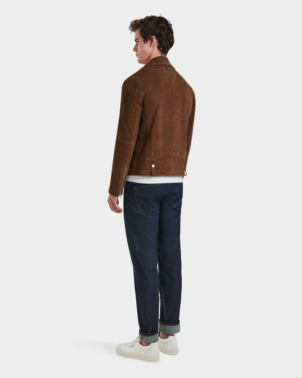 A man in a Rudsak DAREG SU - 6125053 Mocha suede biker jacket, dark blue cuffed jeans, and white sneakers stands against a plain white background, creating a minimalist look.