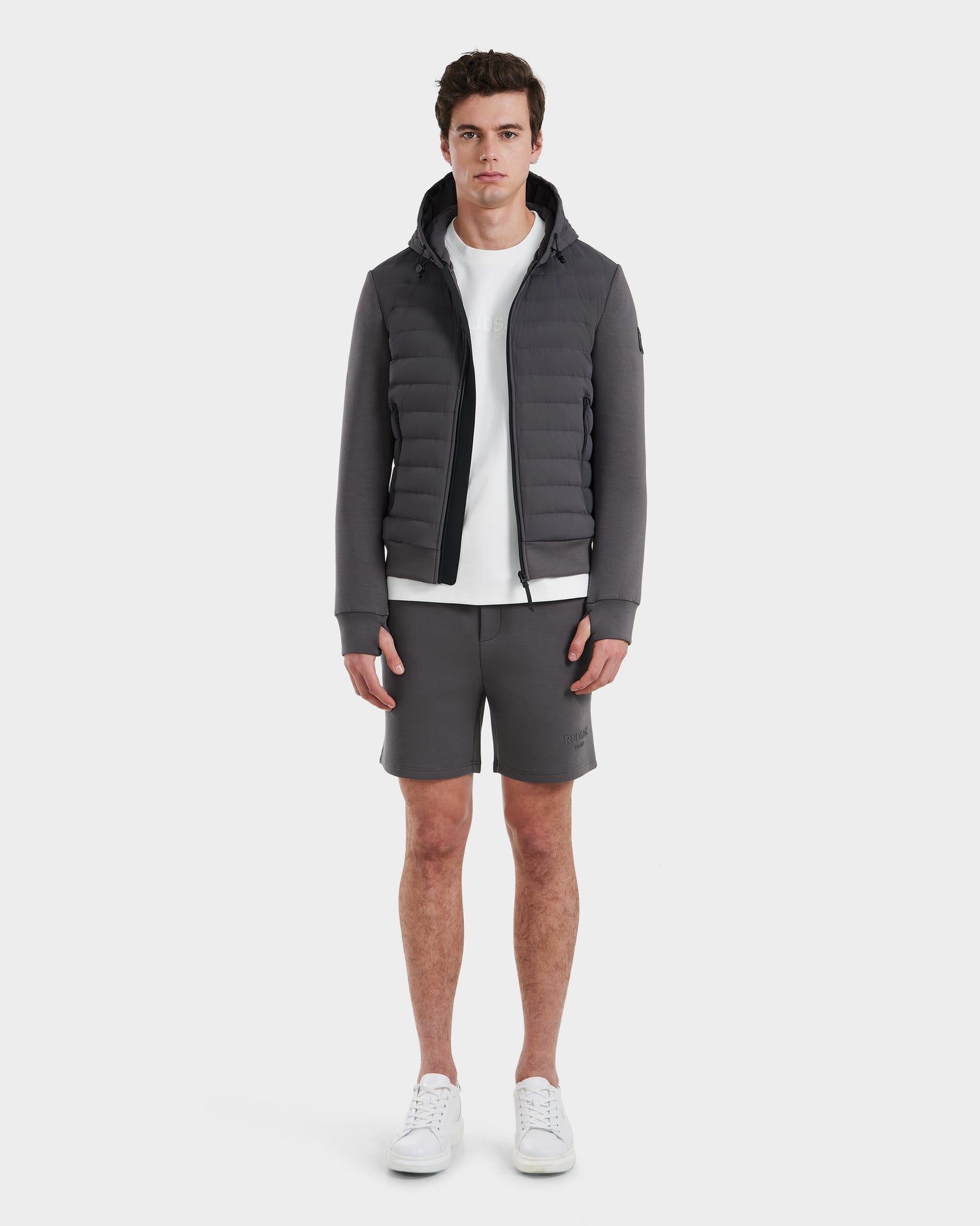 A man faces forward against a plain white background, wearing the Rudsak DEE MAT - 6124028 Grey mens lightweight down jacket, paired with a white t-shirt, gray shorts, and white sneakers.