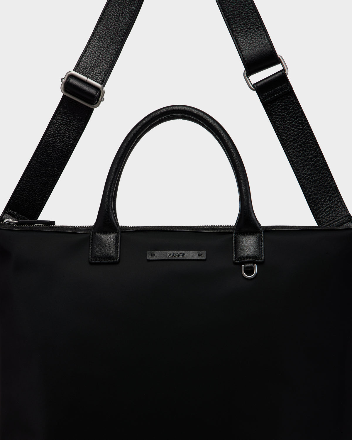 A close-up of the Rudsak DEXTER - 9324513 Black tote, a sleek unisex bag with sturdy leather handles, a detachable shoulder strap, and a small metallic brand label on the front.