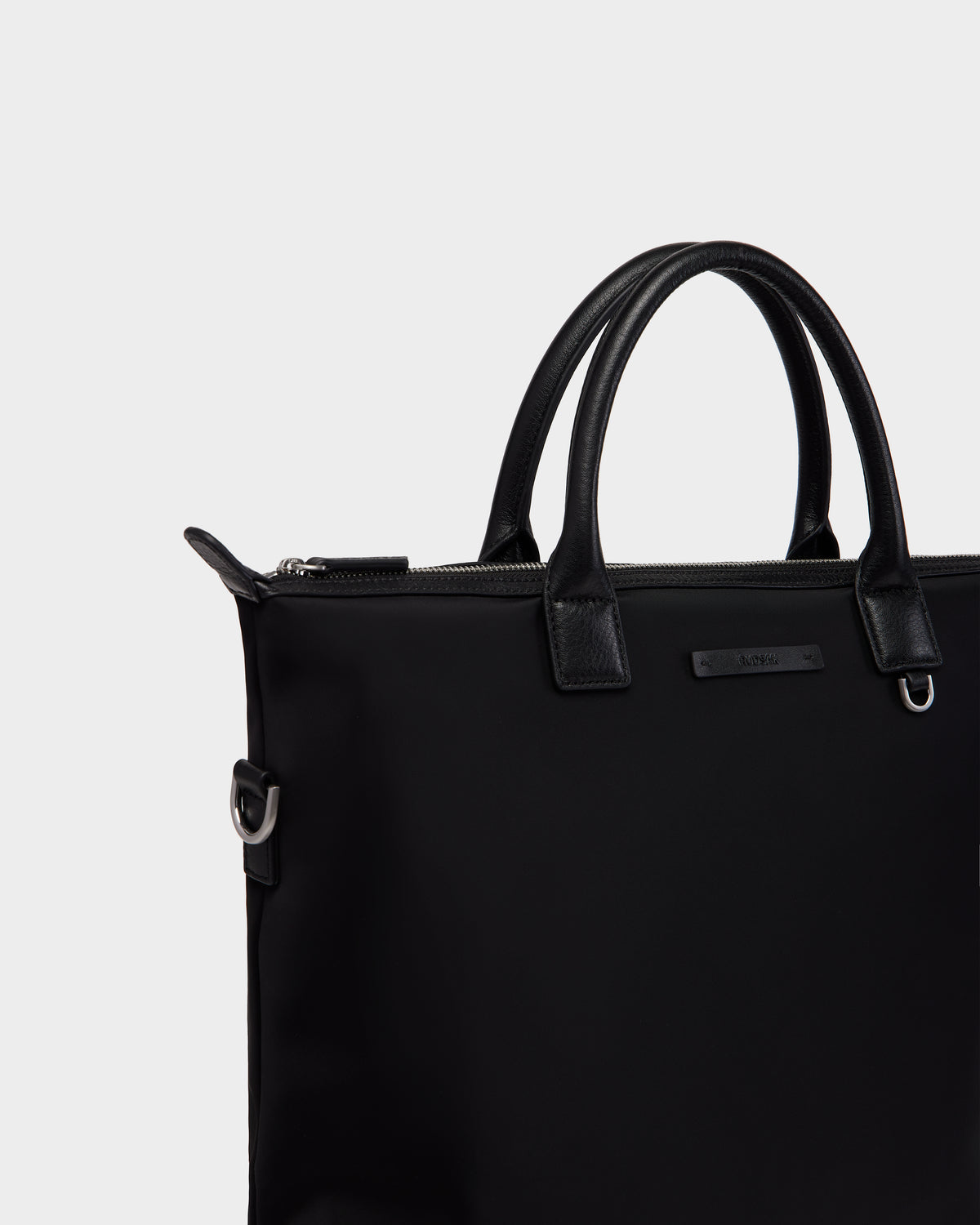 A close-up of the Rudsak DEXTER - 9324513 Black unisex tote bag with leather handles, silver hardware, and a zipper closure, set against a plain light gray background—a stylishly versatile accessory for any occasion.