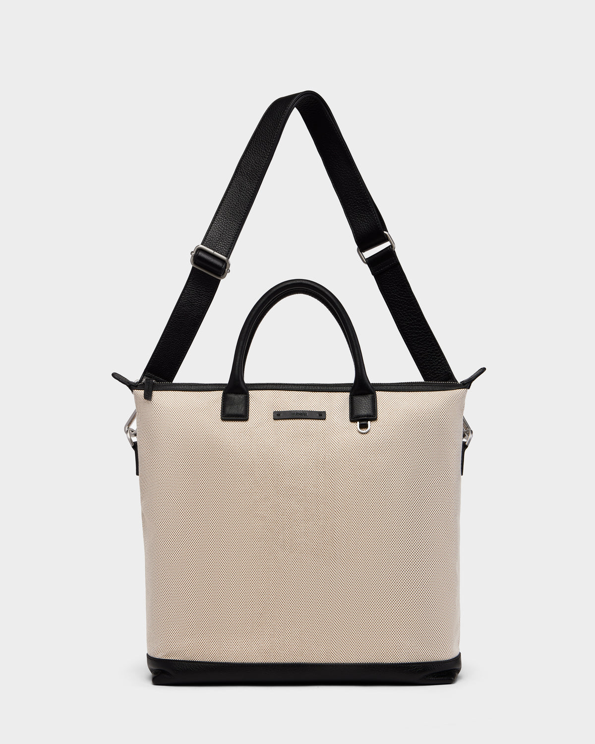 The Rudsak DEXTER - 9324513 Vanilla is a unisex beige canvas tote with black leather handles, a black base, and an adjustable shoulder strap, shown against a plain white background.