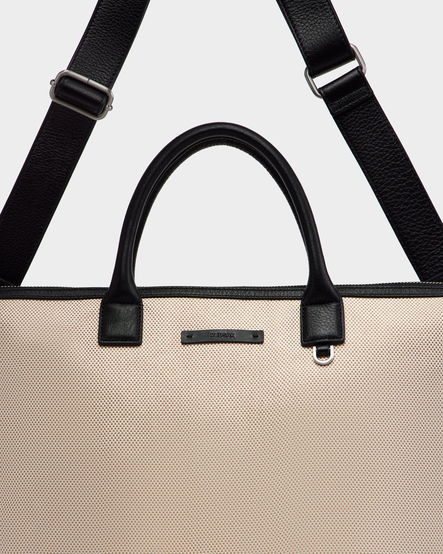 A close-up of the Rudsak DEXTER - 9324513 Vanilla tote bag features beige canvas, black leather handles, and a detachable black shoulder strap, highlighting this versatile accessory against a plain background.