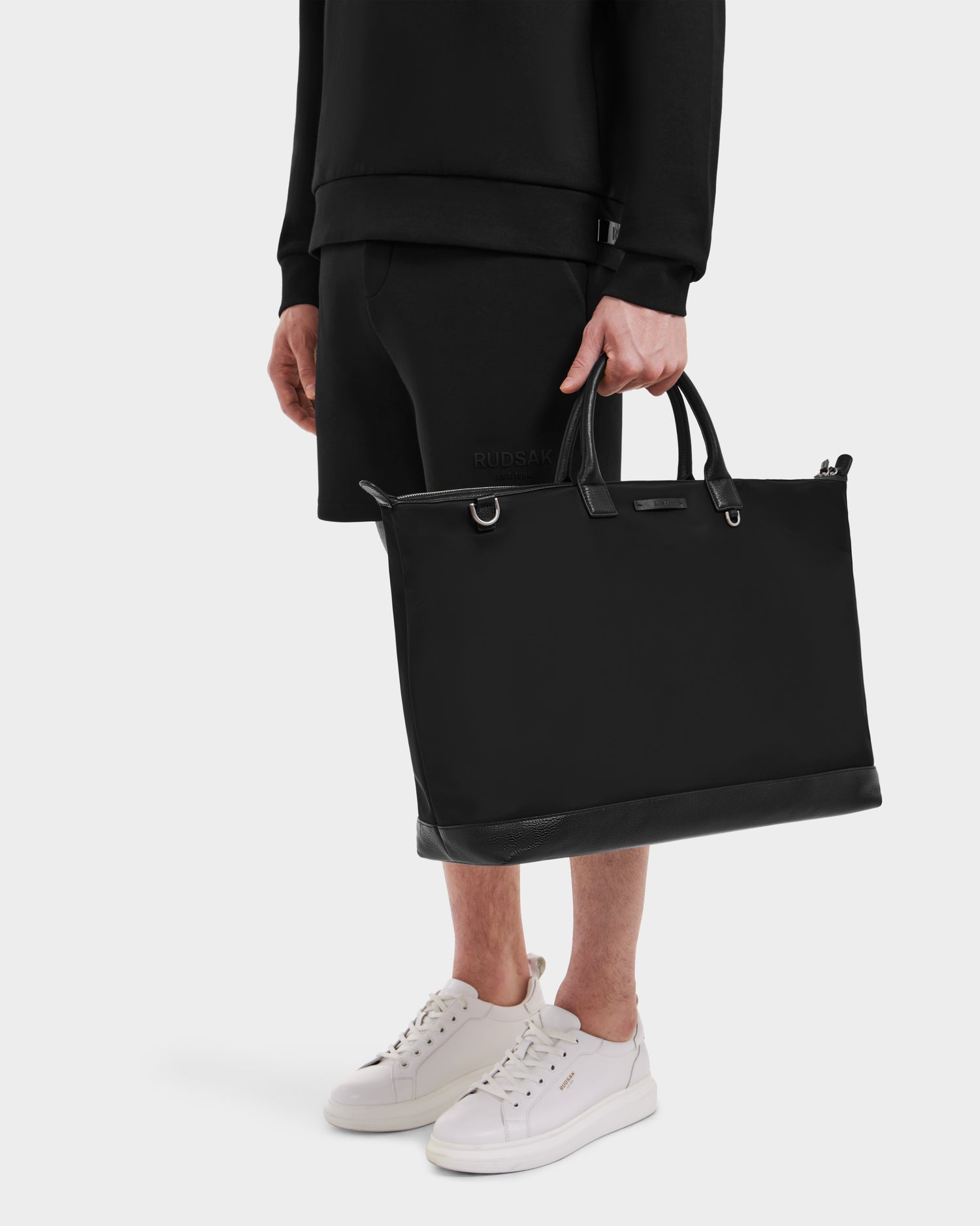 A person holds the Rudsak DUBLIN - 9324514 Black unisex weekender tote by its handles, wearing a black sweatshirt, black shorts, and white sneakers against a plain light background.